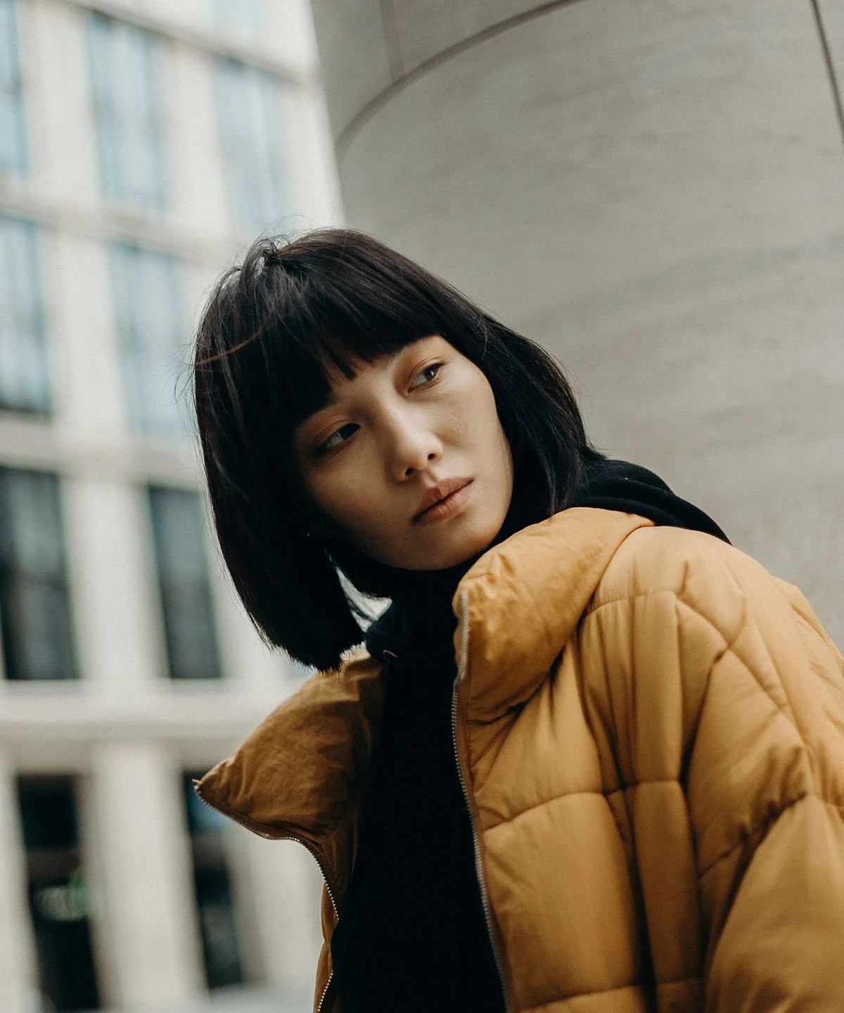 A young woman with black hair and bangs, wearing a tan puffer jacket over a black hoodie, looking to the side with a neutral expression, standing outdoors near a modern building.