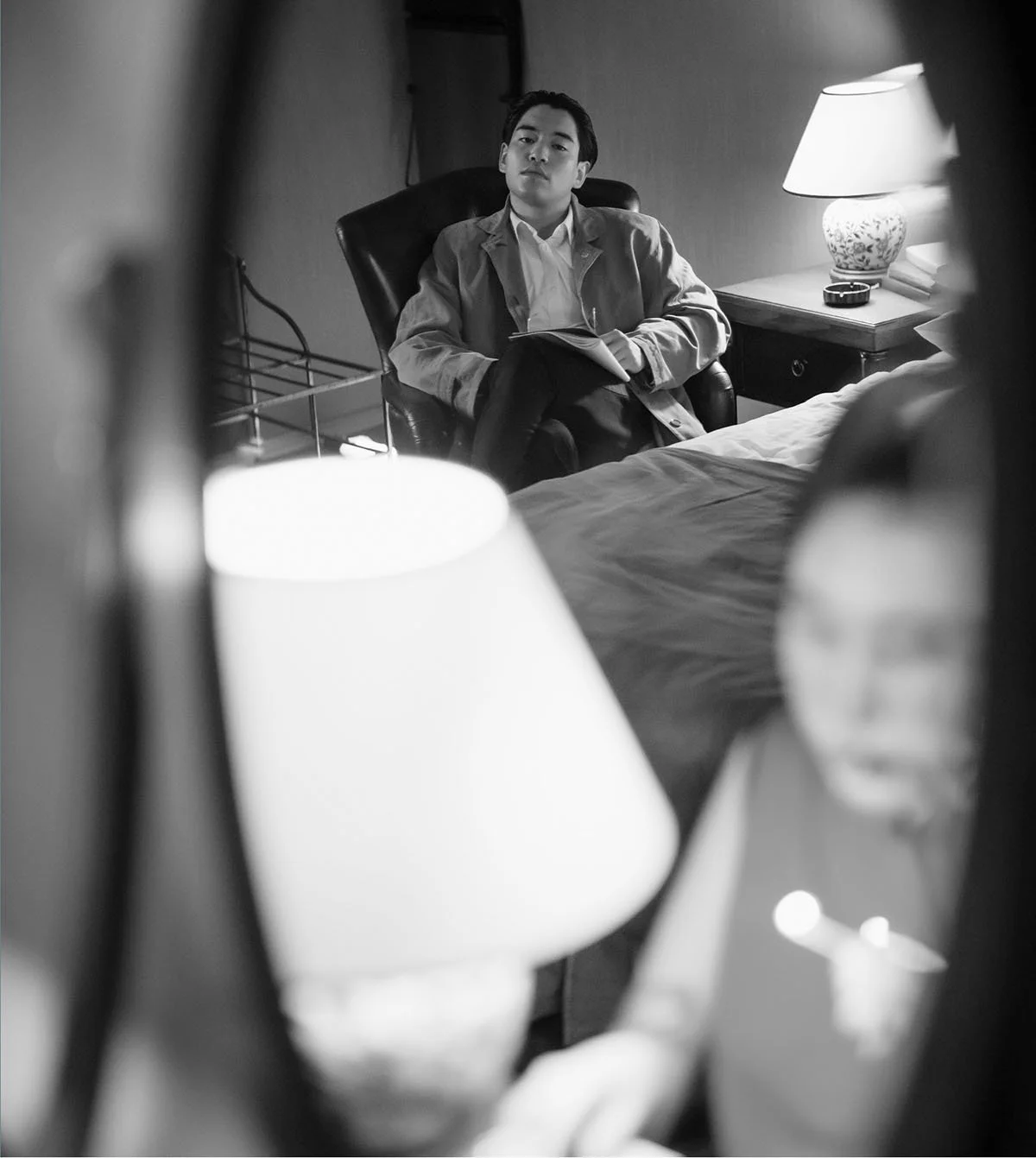 A black and white photo of a man sitting in a room, taken through a mirror. The man appears to be assessing or reflecting, holding a notebook or folder, and is seated in a chair. The room contains a lamp on a bedside table, some books, and a bed in the foreground.