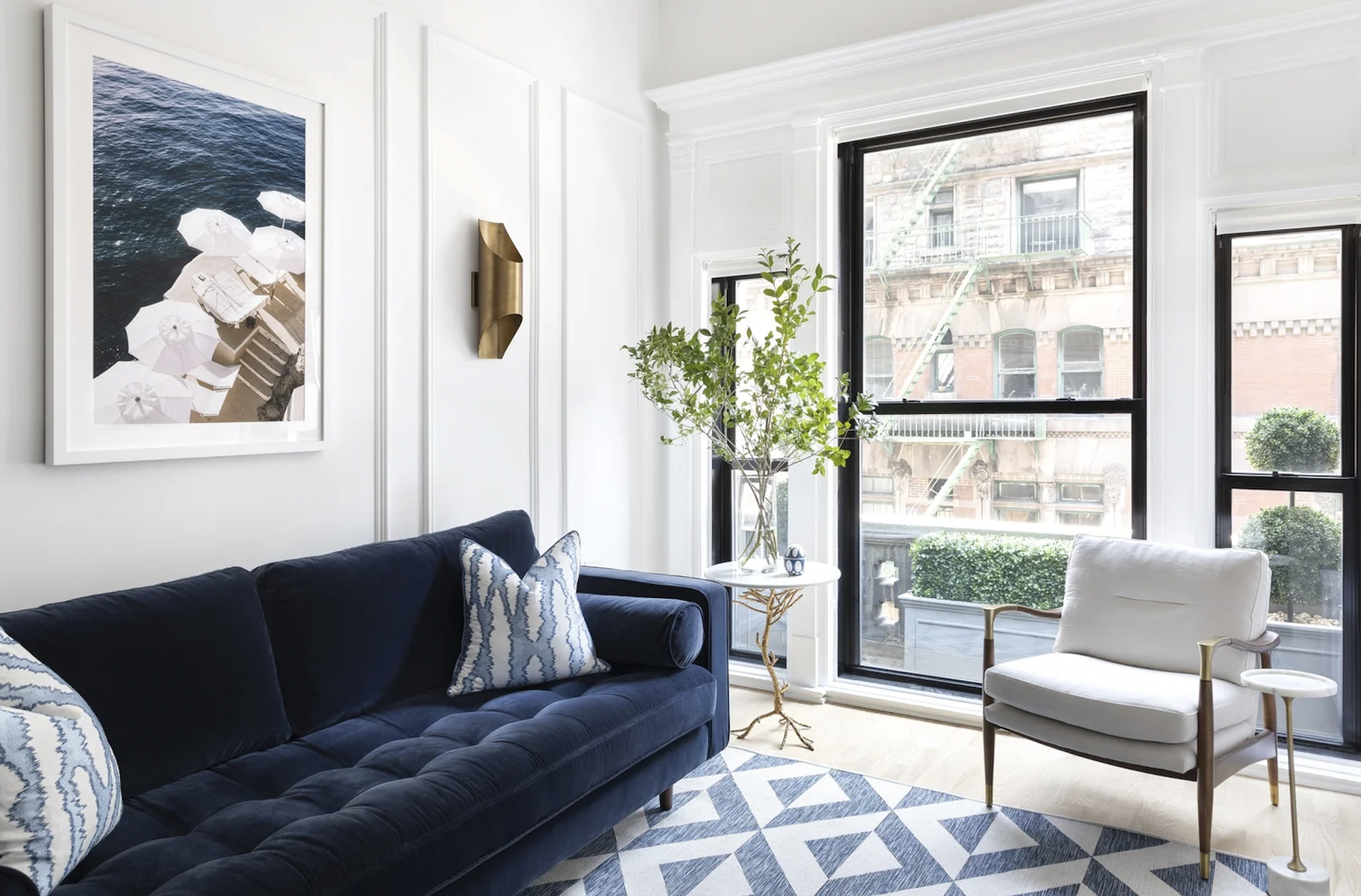 A modern living room with large windows, a navy blue sofa with patterned pillows, a white armchair, a round side table with a glass vase of greenery, framed artwork, and blue and white patterned rug.