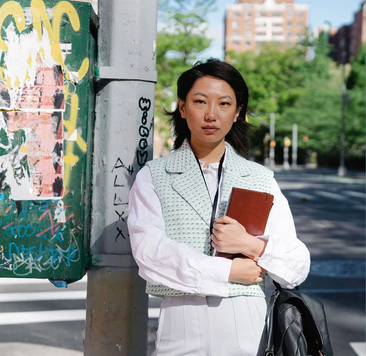 A woman with black hair and light skin standing on a city street holding a brown notebook. She is wearing a white shirt and a light-colored vest, with a black bag hanging on her shoulder. Behind her is a graffiti-covered green electrical box and trees with a building in the distance. The weather appears sunny and bright.