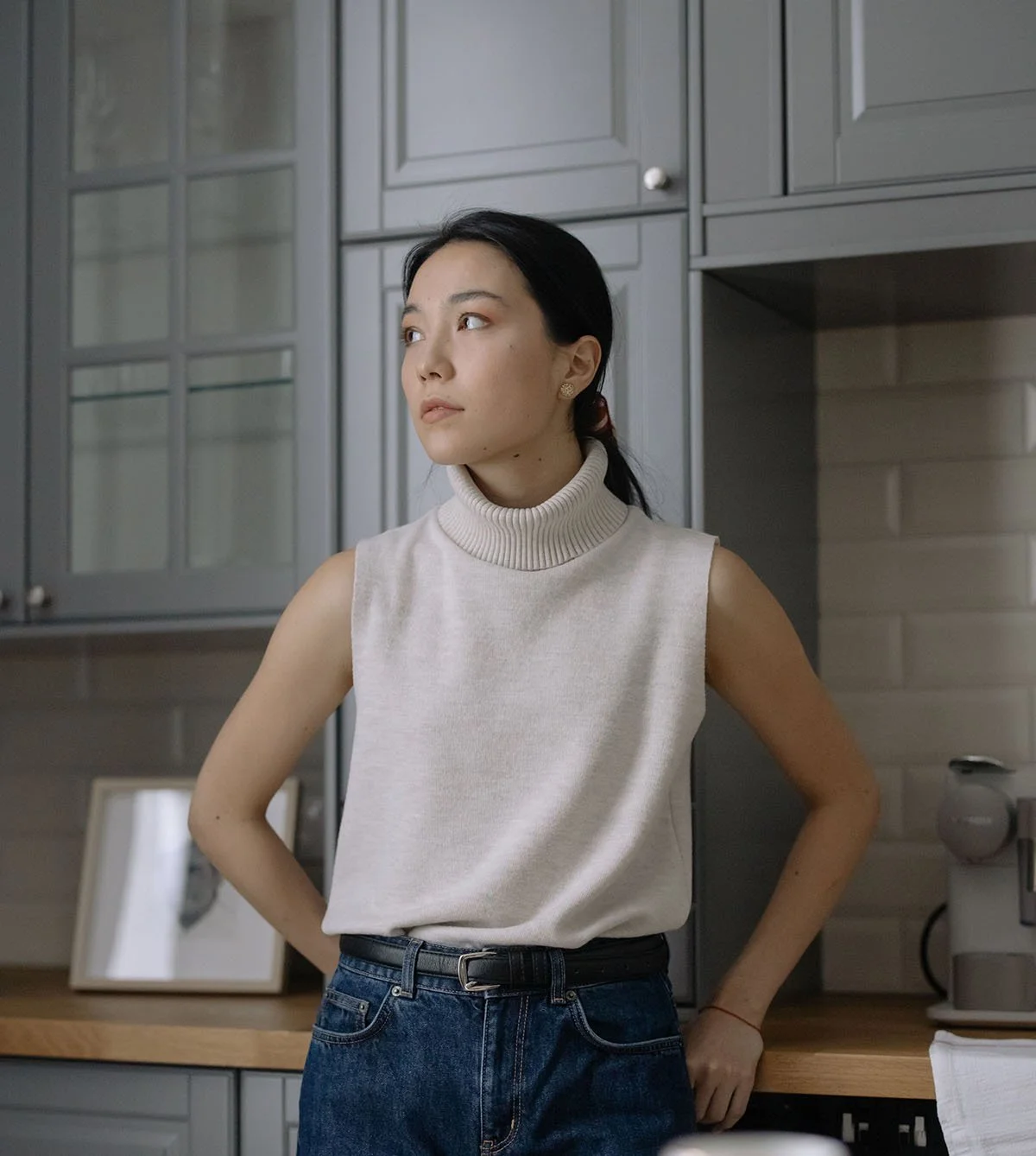 A woman standing in a modern kitchen, looking to her left with her hands on her hips, wearing a sleeveless turtleneck top and jeans.