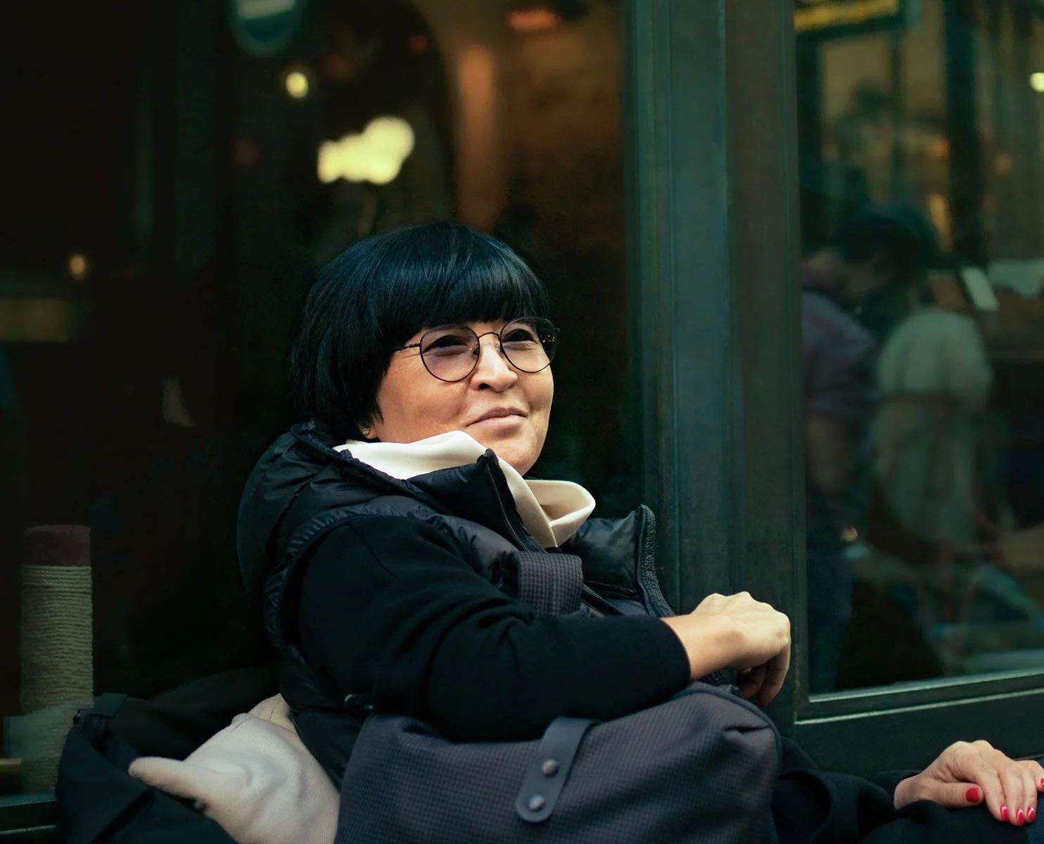A smiling woman with short black hair and glasses, wearing a black jacket and white hoodie, sitting on a bench beside a window.