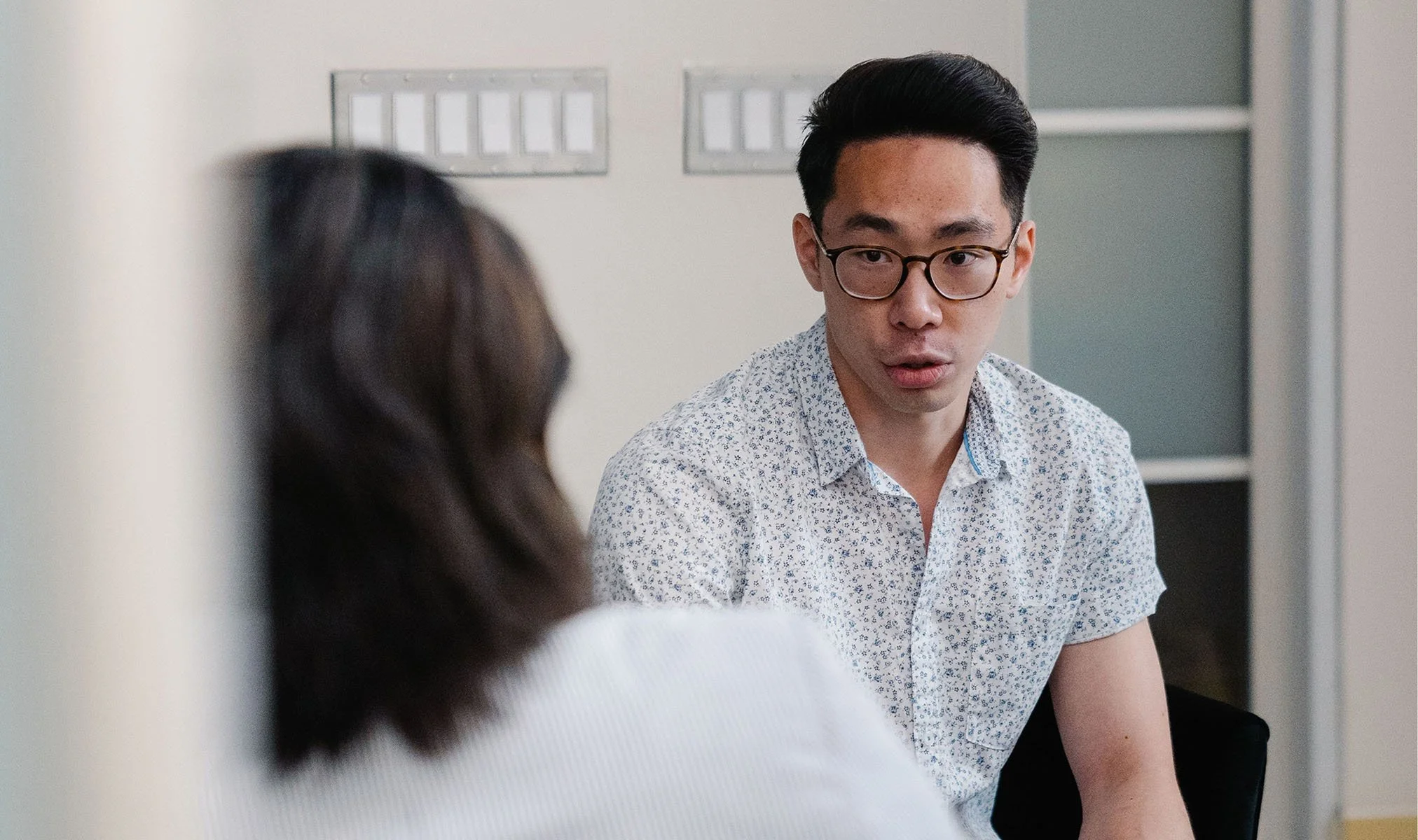 A young man with glasses and a patterned shirt engaged in conversation with a woman who has long dark hair, in an office setting.