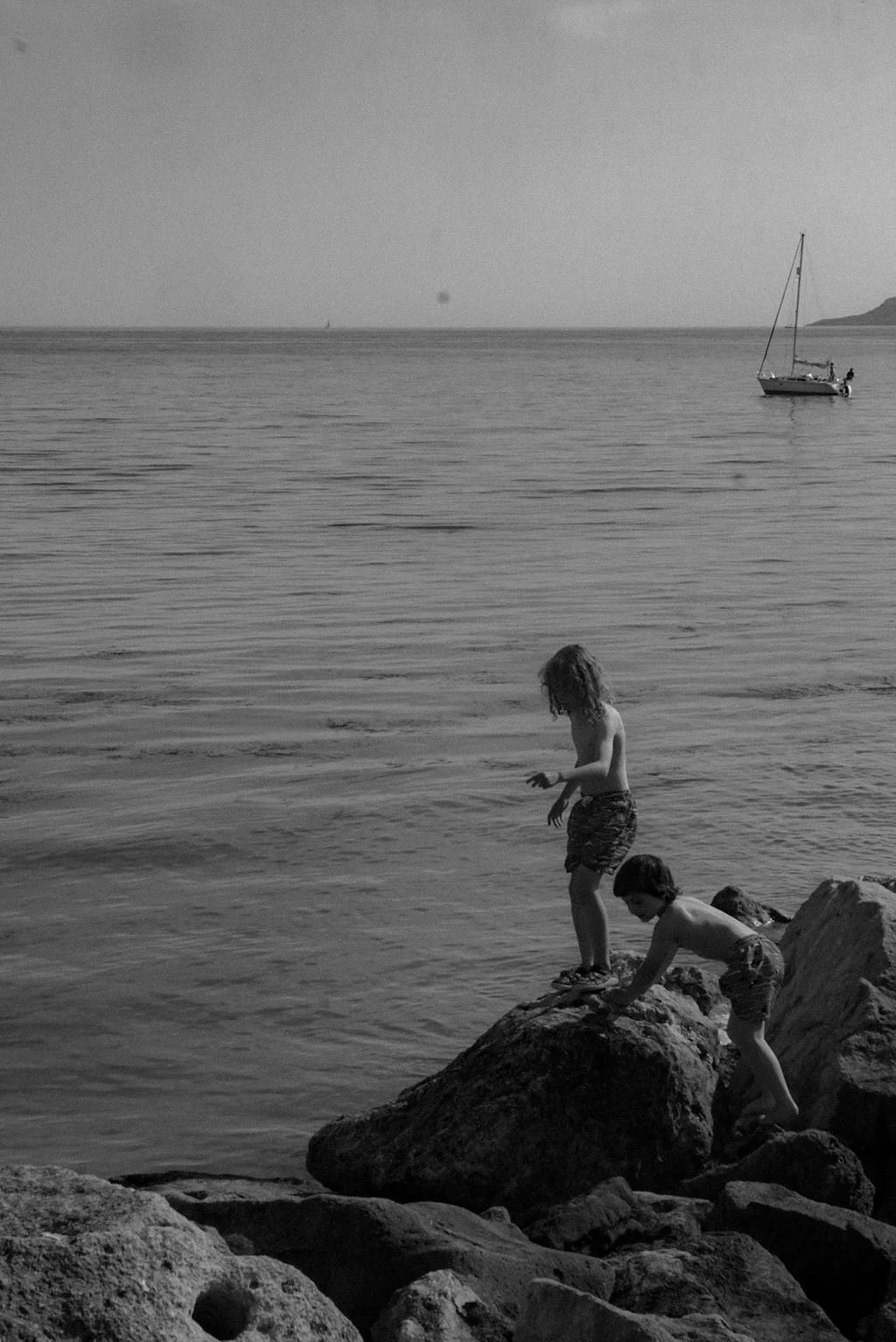Two children playing on rocks by the water, with a boat floating in the distance, on a calm seaside day.