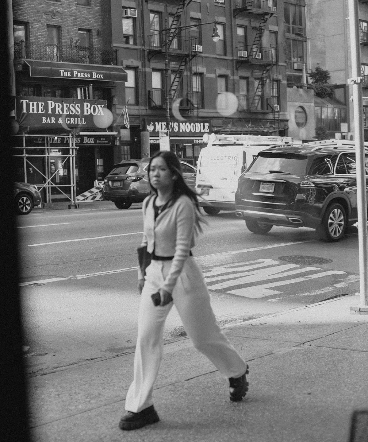Black and white street scene with a woman walking on the sidewalk in foreground, commercial buildings and parked cars in the background, including a sign for The Press Box bar and grill and a restaurant called M E U S Noodle.