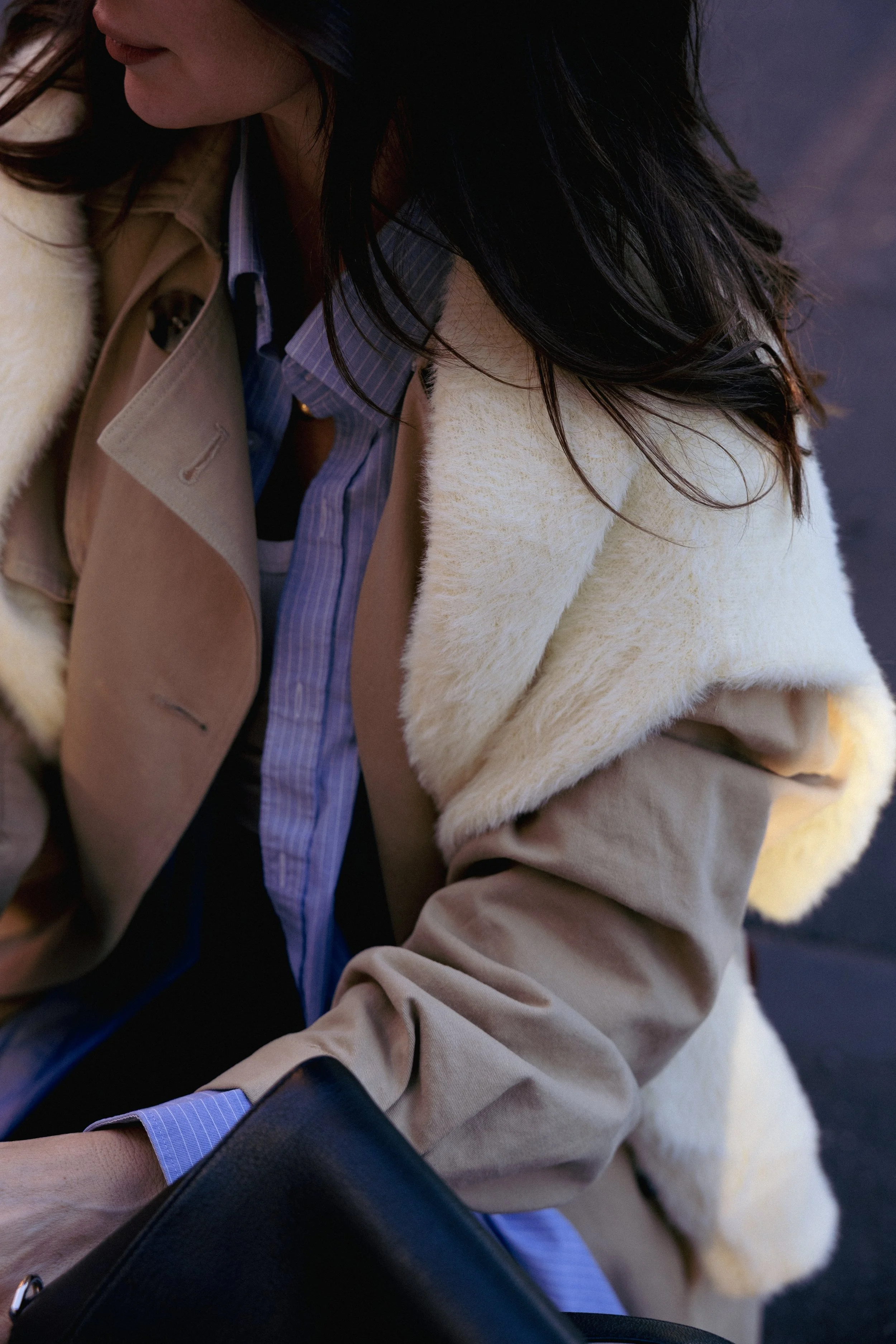 Close-up of a woman in layered fall clothing, including a tan trench coat, cream shearling jacket, and blue button-up shirt, with dark hair, holding a black bag.