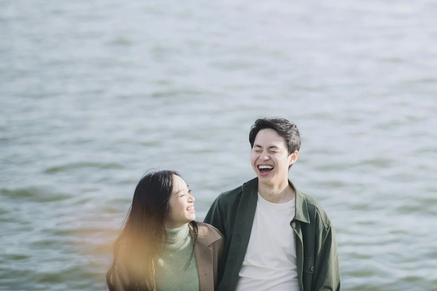 A young man and woman laughing and smiling near a body of water, enjoying a happy moment together.