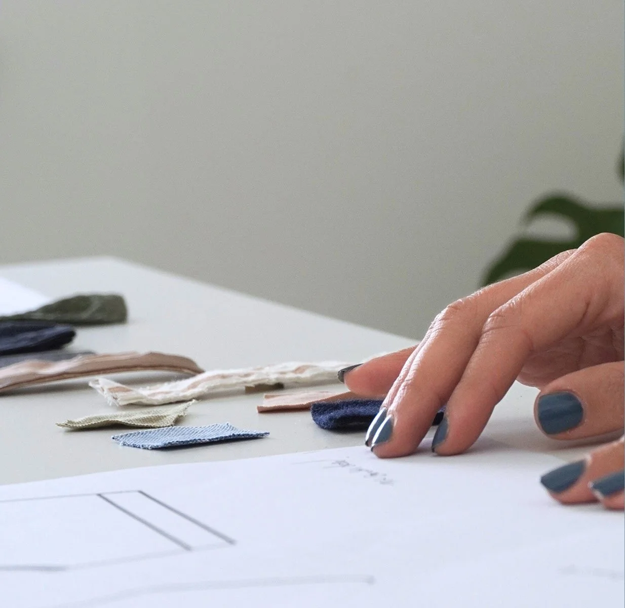 Close-up of a person's hand with painted nails reaching over fabric swatches and design sketches on a white table.