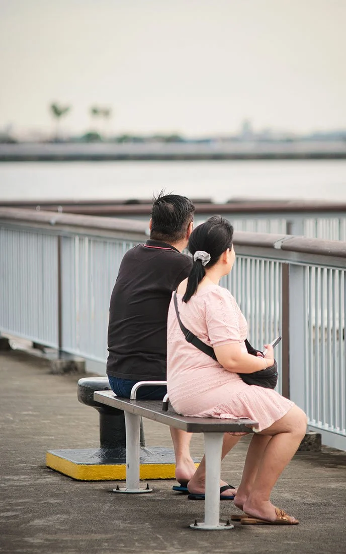 A man and a woman sit on a bench facing a body of water, with a railing and blurry trees or structures in the background, during daylight.