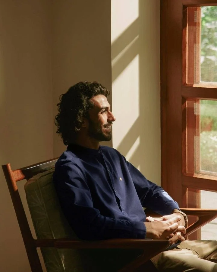 A man with dark curly hair and a beard sitting in a wooden chair near a window, enjoying sunlight and smiling.