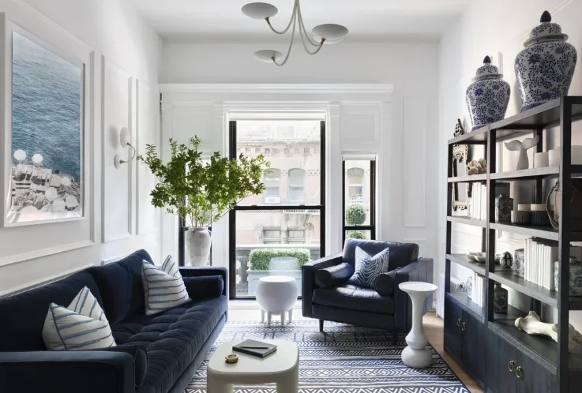 Bright living room with black sofa and armchair, white walls with decorative molding, large window with city view, black bookshelf with decorative items, potted plant, striped rug, and white side tables.