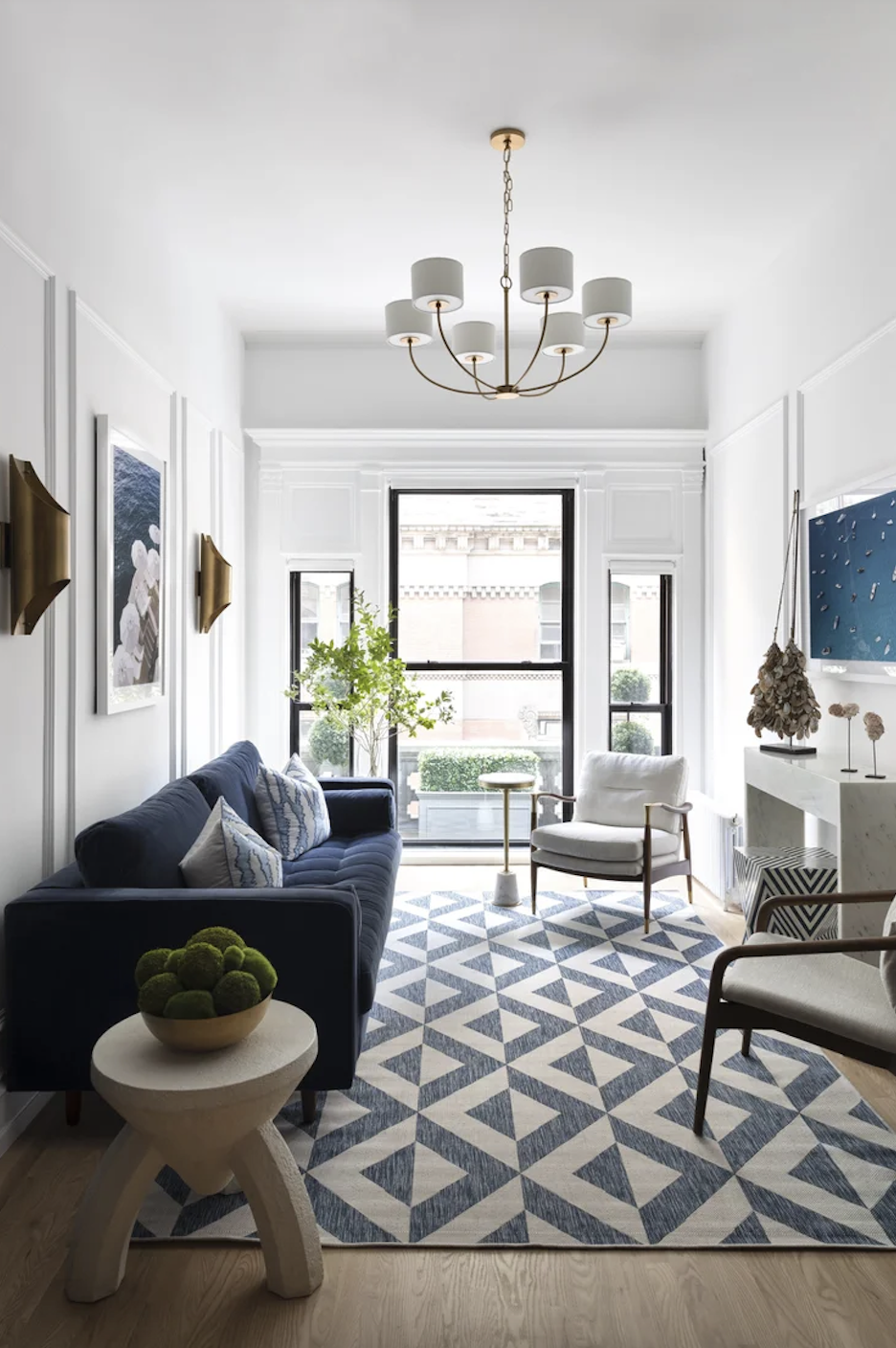 Modern living room with white walls, a navy blue sofa, a white armchair, and a geometric-patterned rug. Large windows let in natural light, and a chandelier hangs from the ceiling. Decorative elements include wall art, a small side table, and a bowl 