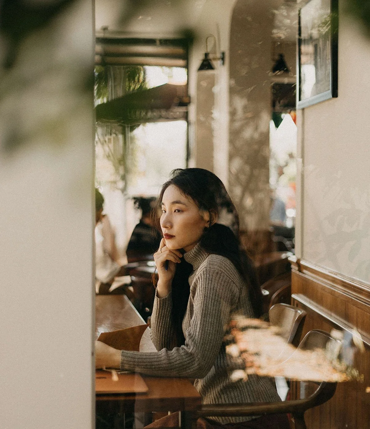 A young woman with dark hair and a beige ribbed turtleneck sweater sitting at a wooden table inside a cozy cafe, lost in thought with her hand resting on her chin, surrounded by blurred figures of other patrons and warm, natural lighting.