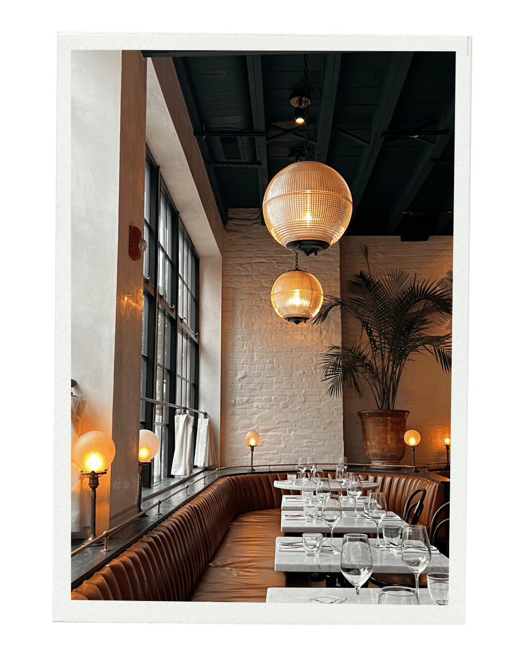 Elegant restaurant dining area with white marble tables, glassware, leather banquette seating, large potted plant, and hanging pendant lights in warm yellow glow, featuring large windows and ambient lighting.