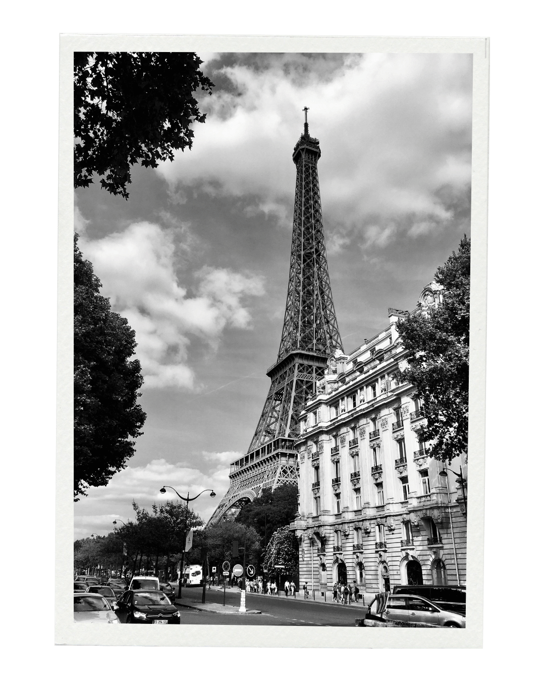 Black and white photo of the Eiffel Tower viewed from the street with cars and pedestrians, trees, and classic Parisian buildings in the foreground.