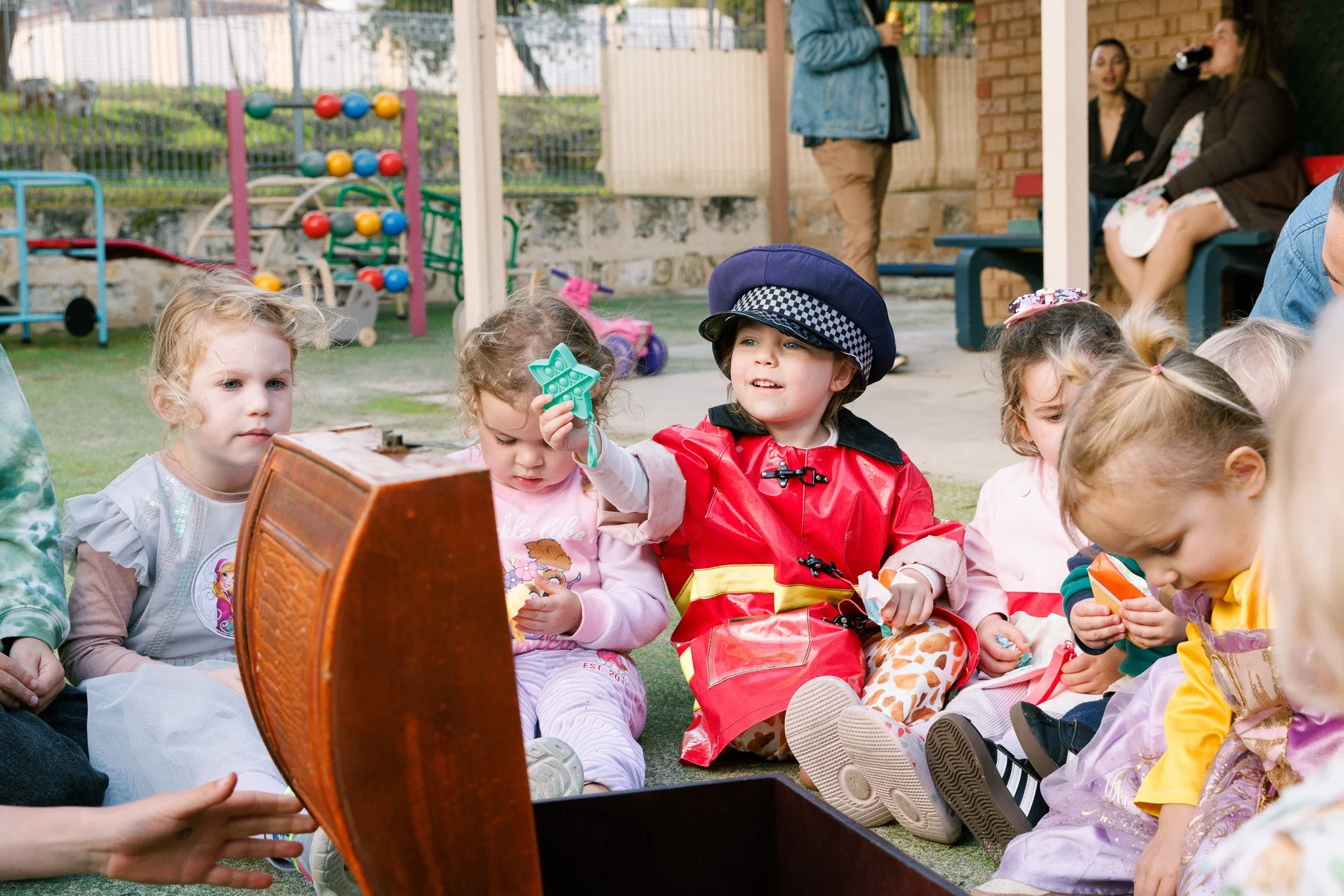 Children sitting outdoors on the ground at a birthday party, with toys and a small wooden chest in front. Some children are dressed in colorful clothes, one with a police hat and red raincoat, others are engaging with toys. In the background, there a