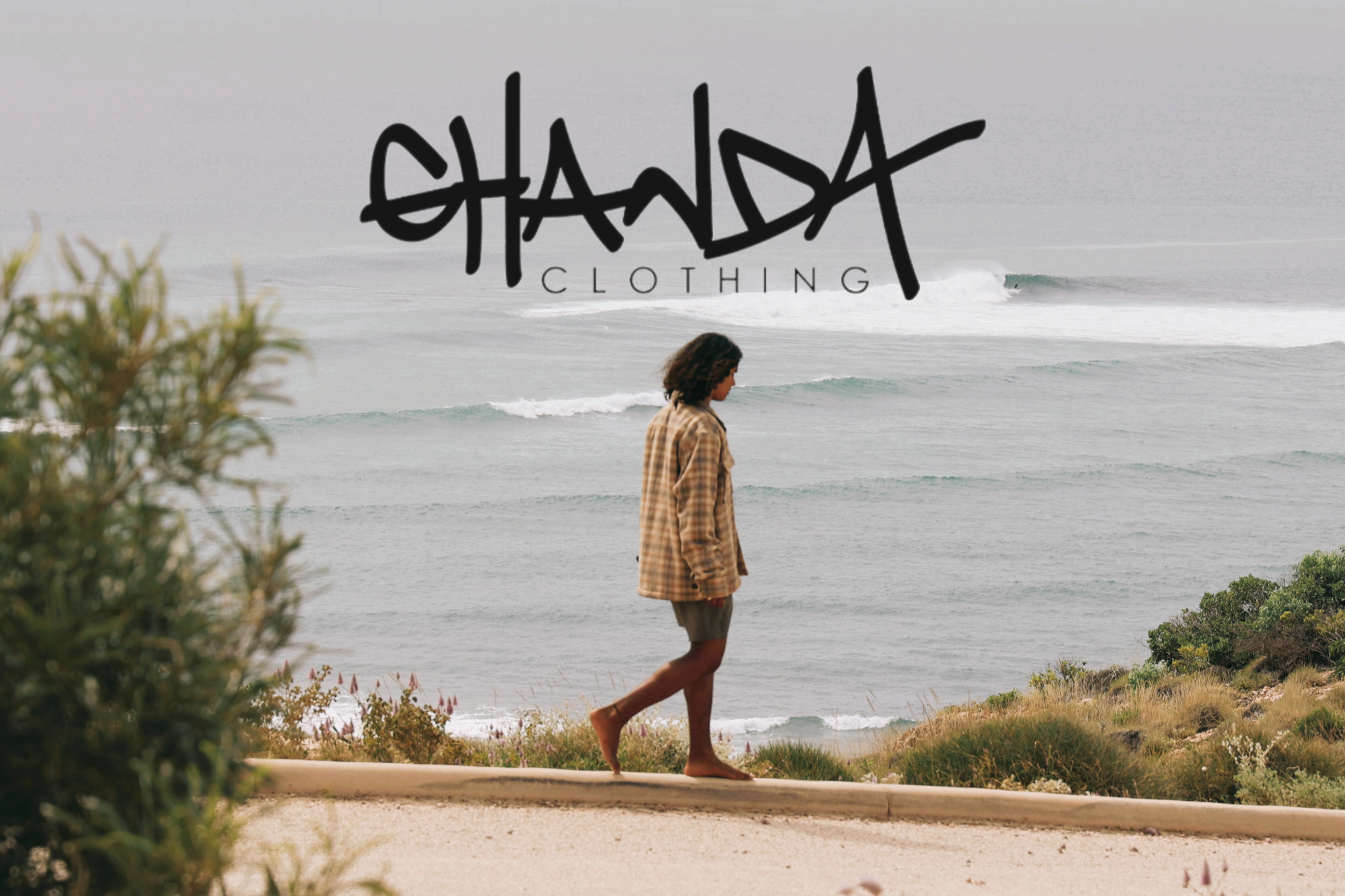 A person walking along a grassy path near the beach, with the ocean waves and surfer in the background and the logo 'SHANDY Clothing' in bold black letters across the center.