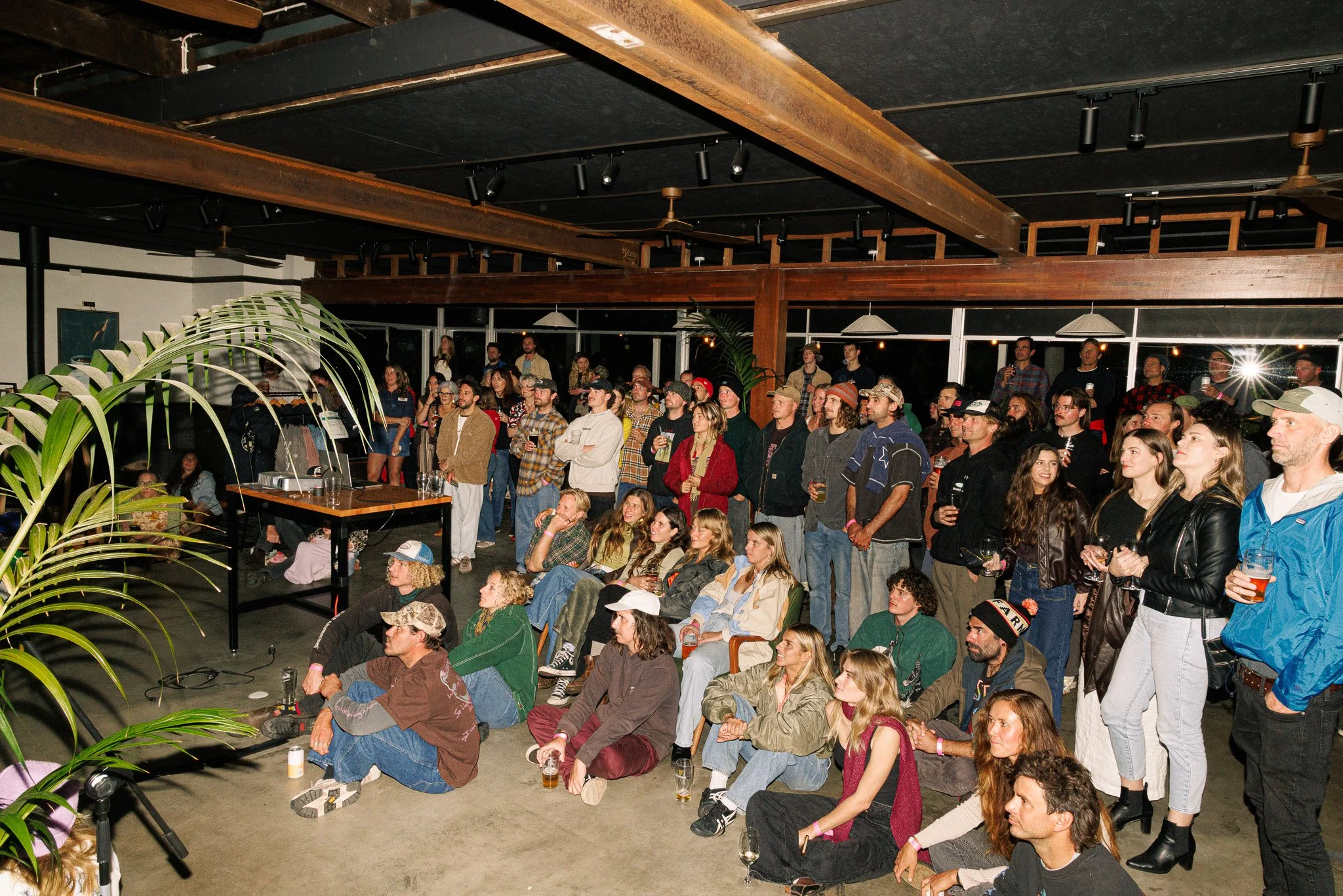 A large crowd of people attending an indoor event, with some standing and others sitting on the floor. The room has a black ceiling with wooden beams, large windows in the background, and some plants in the foreground. Some people are holding drinks,