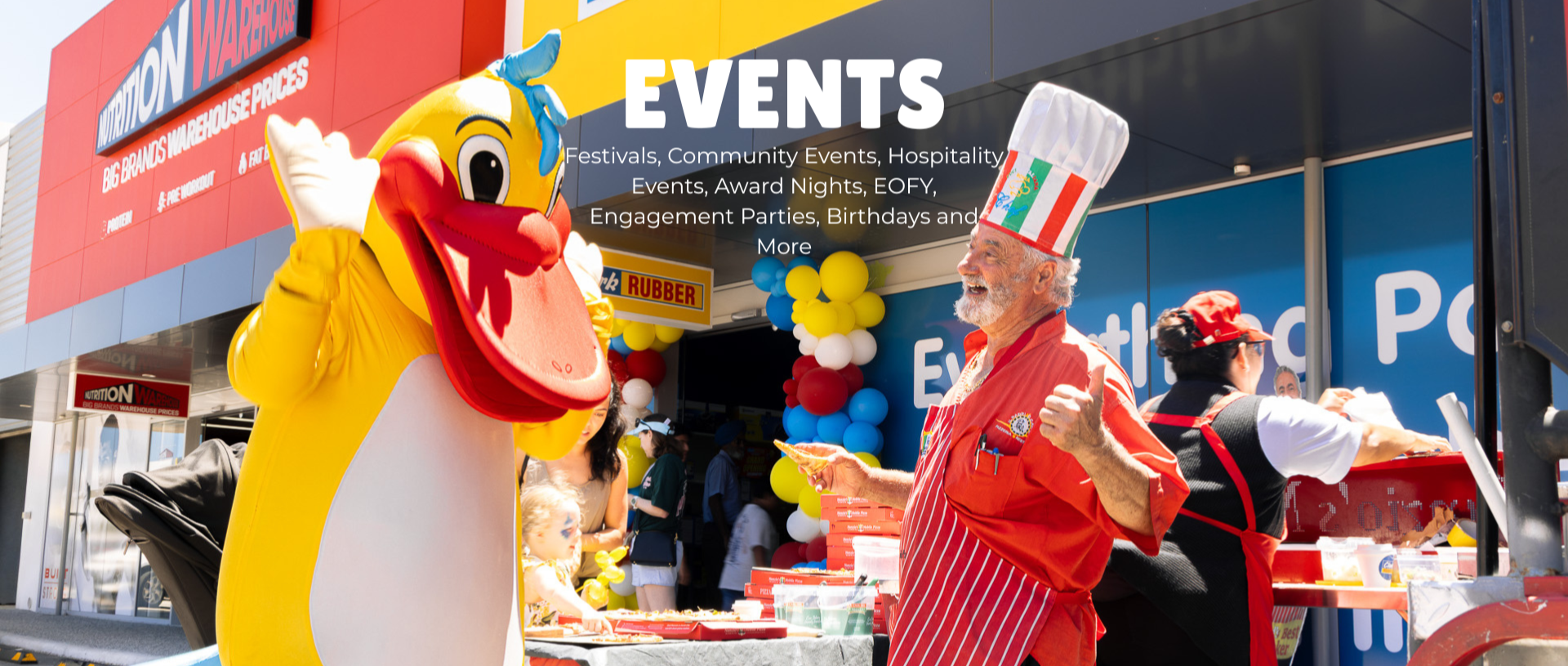 A person dressed in a large chicken mascot costume with bright yellow feathers and a big beak, waving at an outdoor event. An elderly man wearing a red apron and a chef's hat with colorful stripes is smiling and holding a slice of pizza. There is a table with pizza boxes and a woman behind it, serving food. The background shows a building with balloons and promotional signs, indicating a community or festival event.