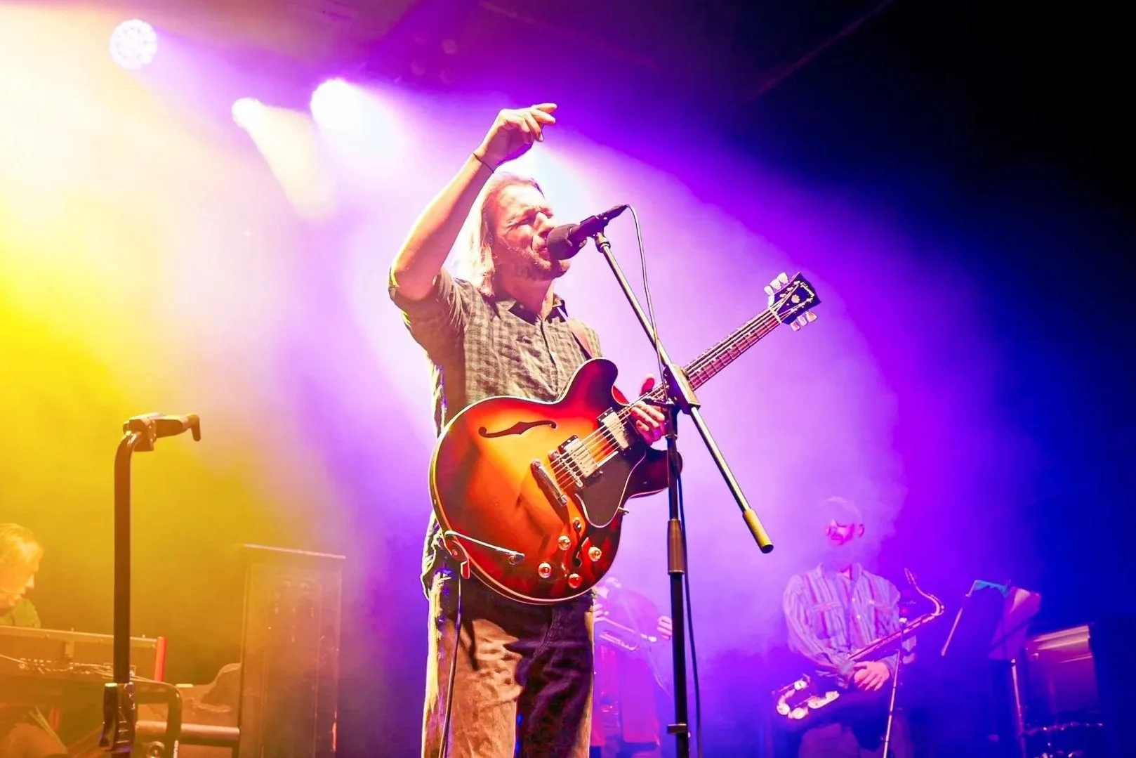 A musician with long blonde hair and beard plays an acoustic guitar on stage, singing into a microphone with purple and yellow stage lights illuminating him.