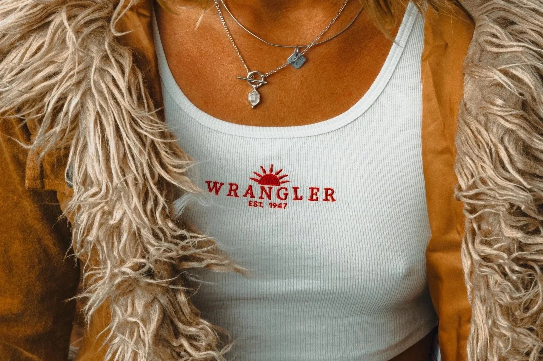 Close-up of a woman's chest and upper torso showing layered necklaces, a Wrangler t-shirt with red embroidery, and a brown jacket with a faux fur collar. She is sitting against a brown background.