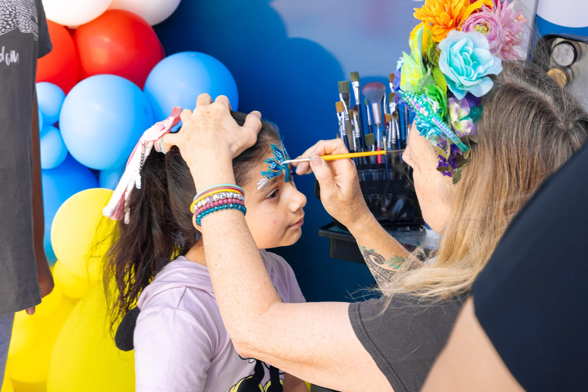 A woman with colorful flowers in her hair face painting a young girl at a festive event with balloons in the background.