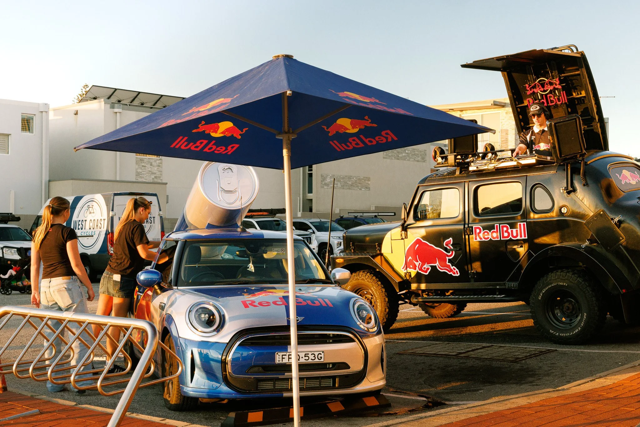 A Red Bull racing theme setup with a small silver car and a larger off-road vehicle behind it, both featuring Red Bull branding. There is a large Red Bull umbrella providing shade, and a person wearing a Red Bull shirt is on the off-road vehicle. Two women are walking past, and a parking lot with white buildings is in the background.