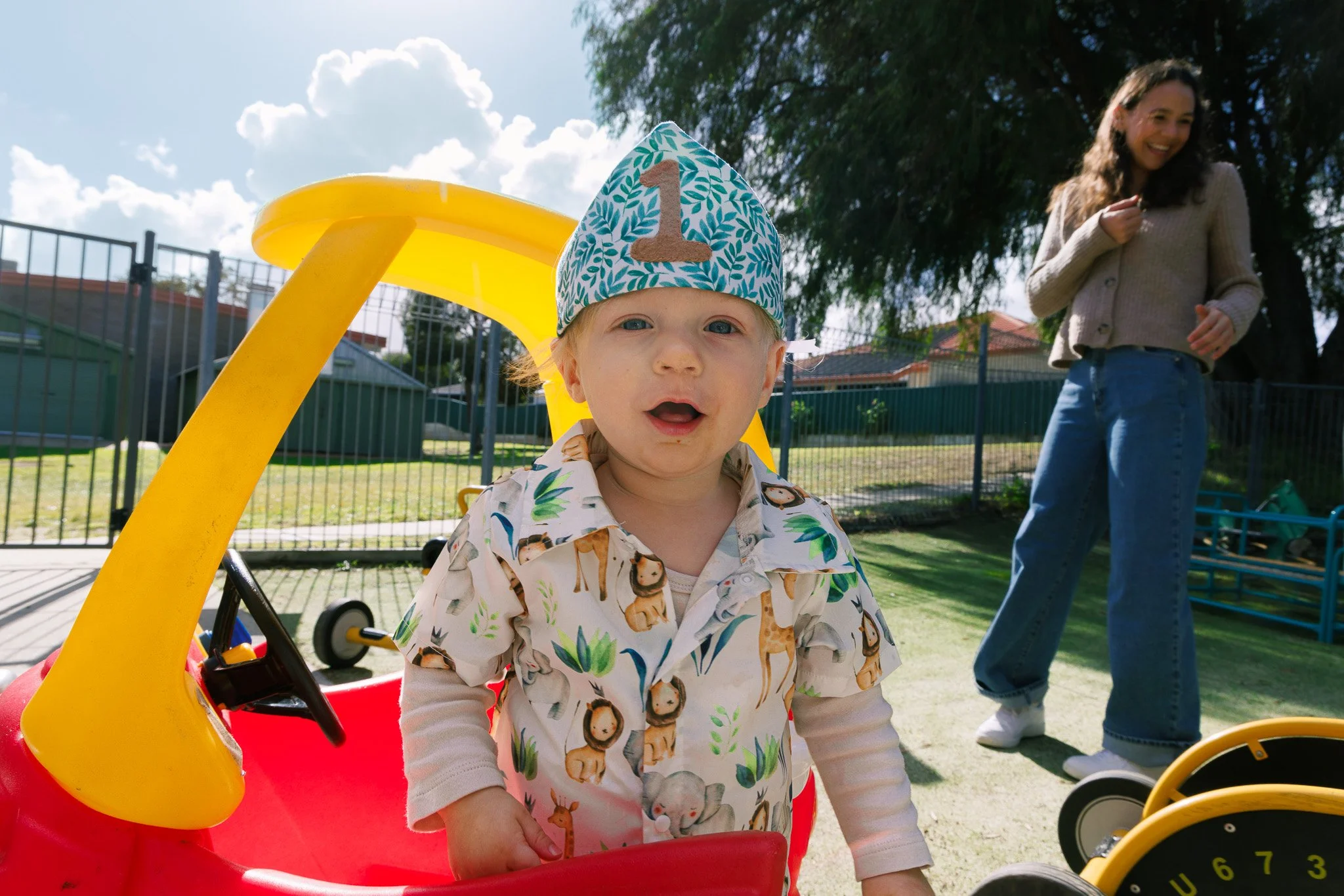 A young boy with blonde hair and blue eyes wearing a colorful safari-themed shirt and a birthday hat with the number 1, standing outdoors near a yellow and red plastic toy vehicle, with a woman smiling in the background beside a large tree and a blue fence.