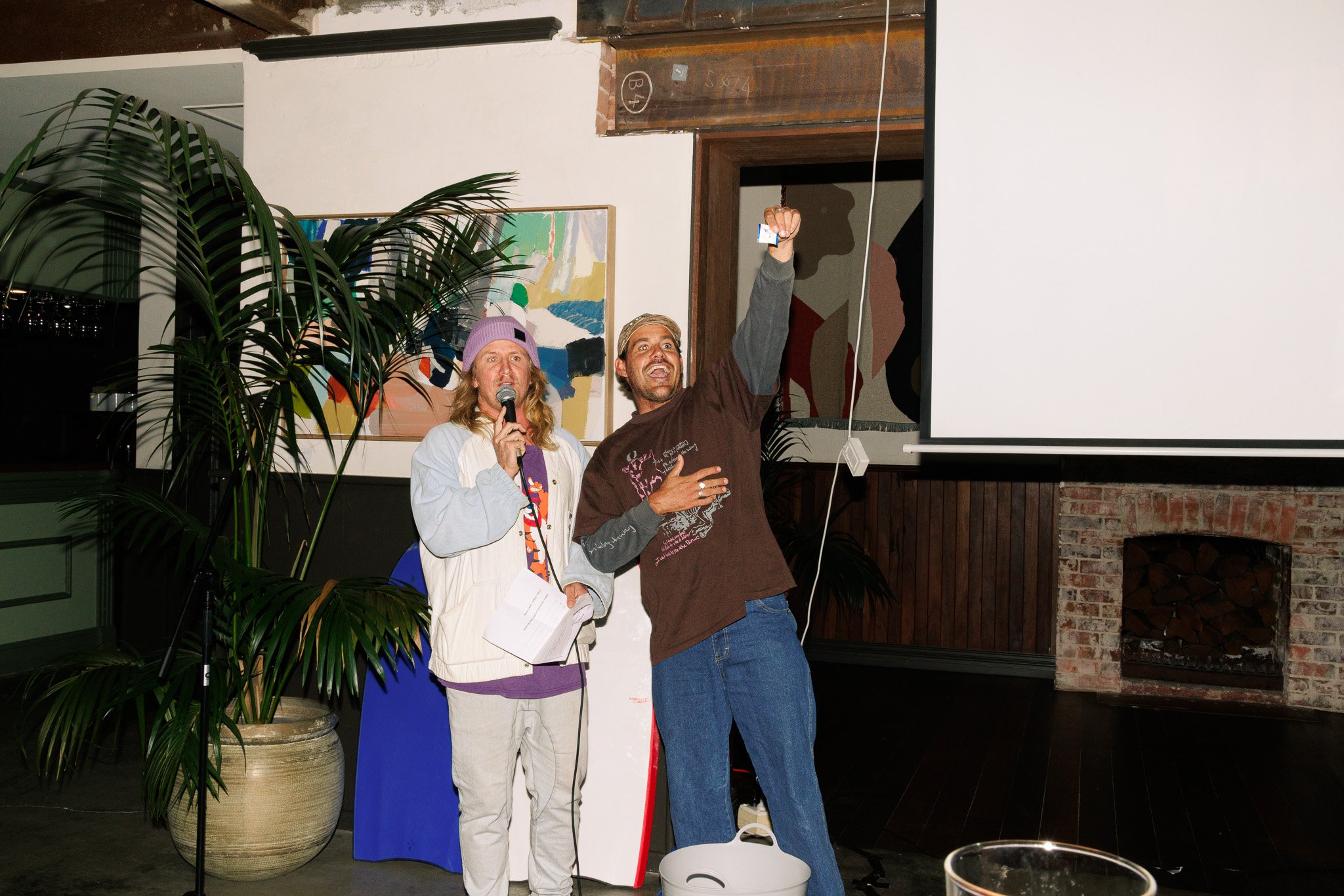 Two people standing together at an indoor event. The woman on the left is holding a microphone and wearing a purple beanie, white and purple jacket, and beige pants. The man on the right is smiling, holding a phone in the air, and wearing a brown T-s