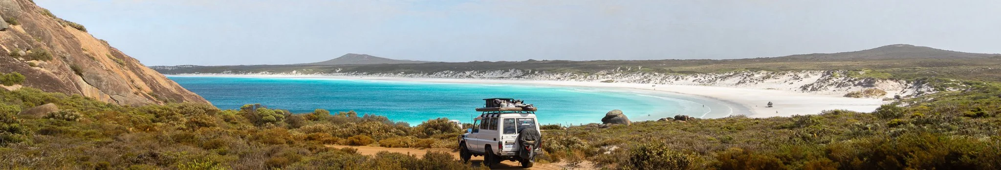 A white SUV parked on a dirt road overlooking a beautiful beach with turquoise water and white sand, surrounded by green and rocky terrain.