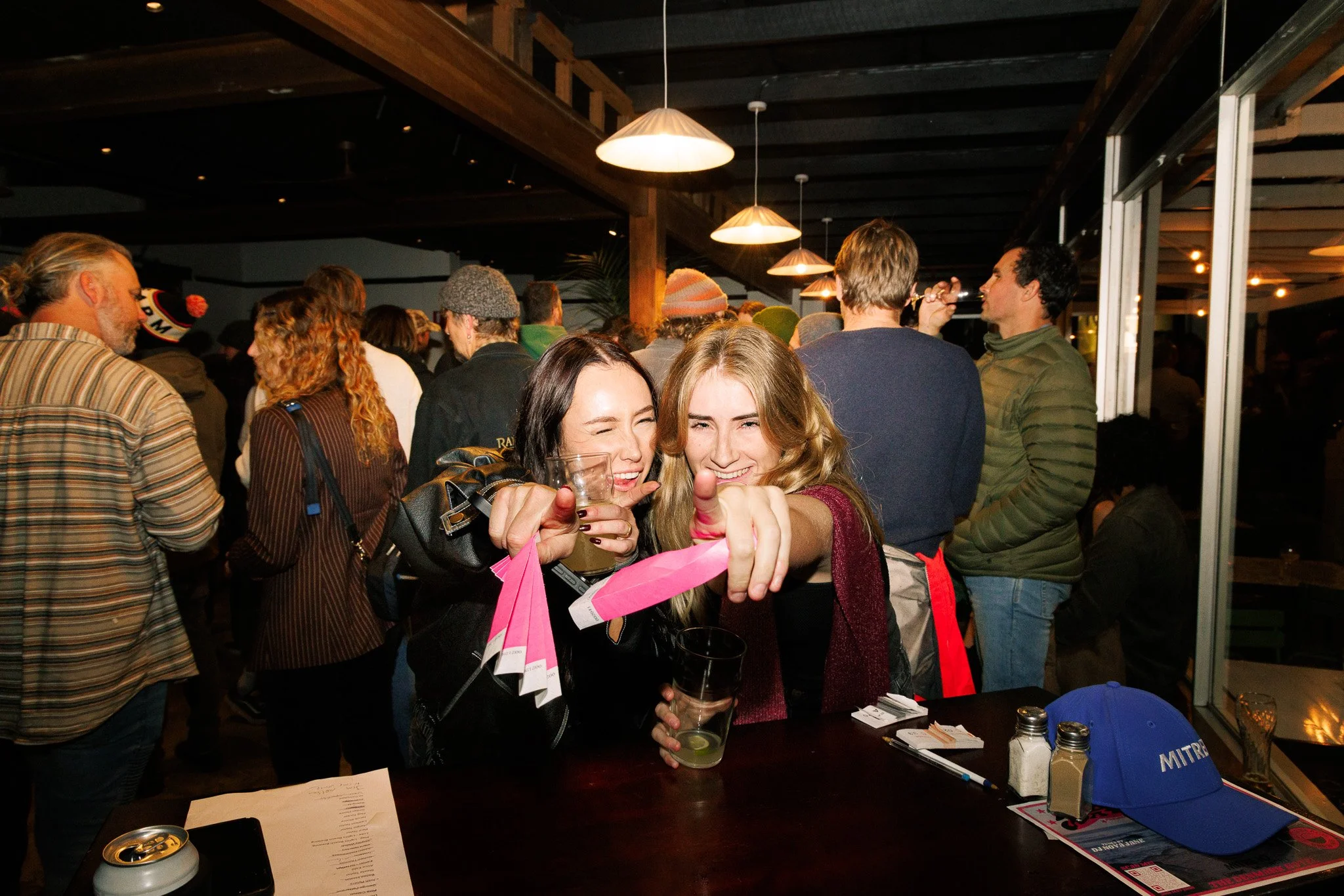 Two young women smiling and taking a selfie in a crowded indoor event space. One is holding a cocktail glass and a pink shopping bag, and they are standing at a table with caps, papers, and drink bottles on it.