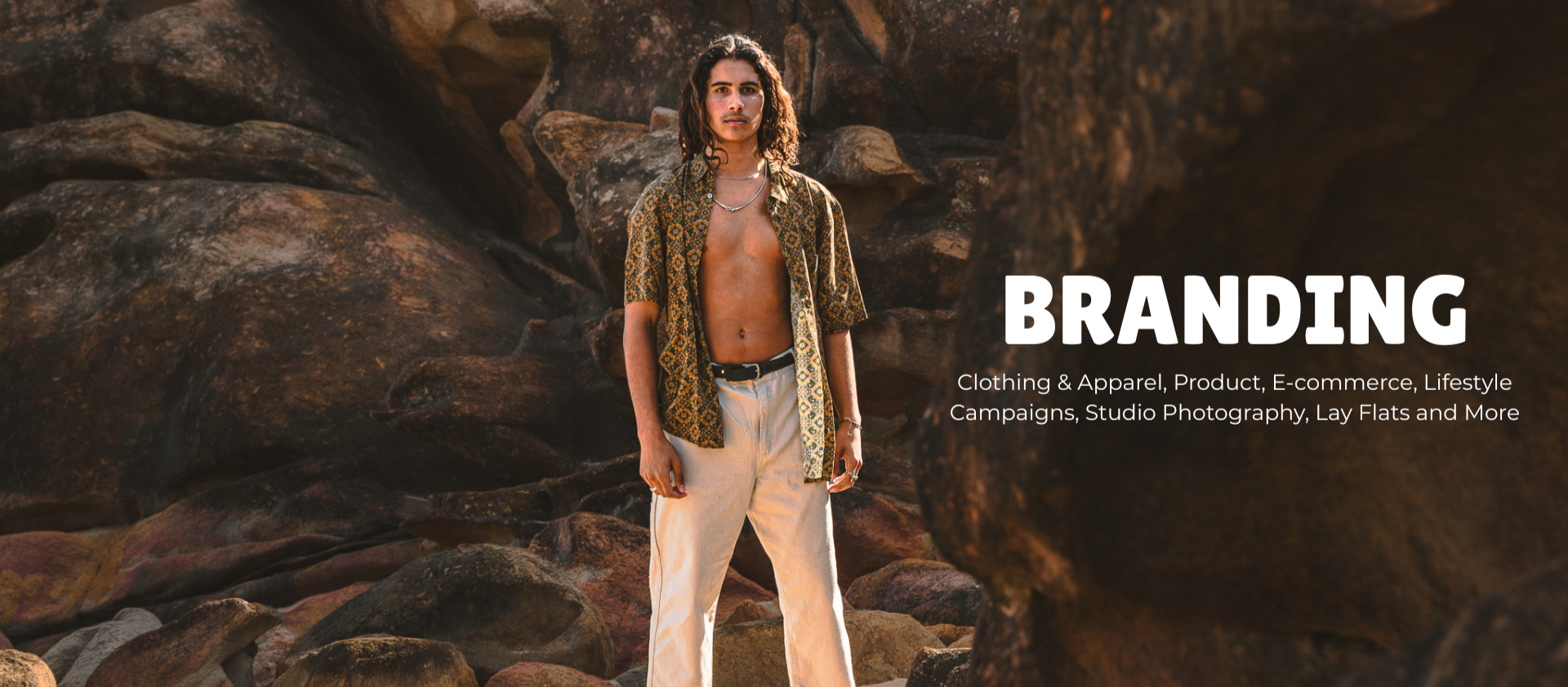 A young man with long, wavy hair stands among rocks in a desert landscape, wearing an open patterned shirt and beige pants, with a necklace, showcasing a fashion and branding theme.