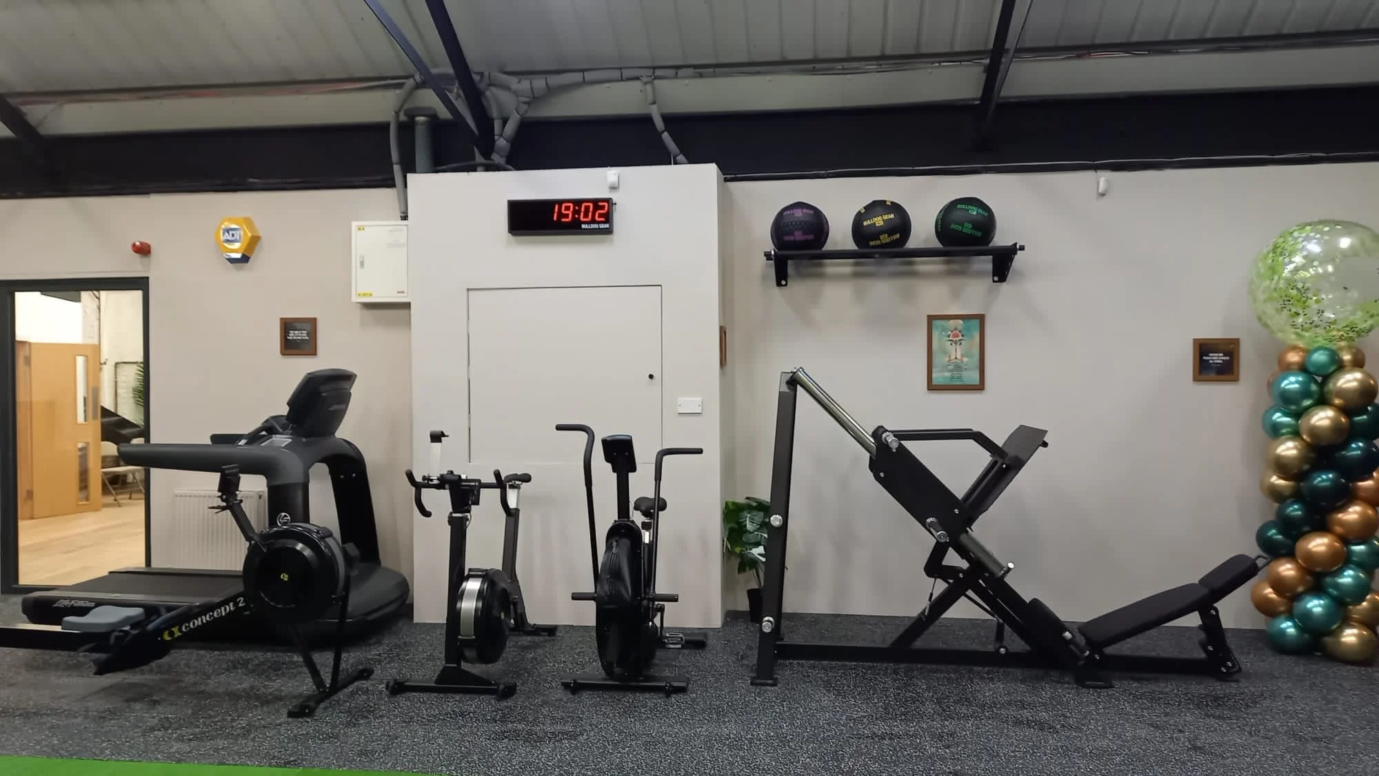Gym interior with exercise equipment including treadmill, stationary bikes, weight machine; wall clock showing 19:02; medicine balls on shelves; framed pictures and balloon decorations. TRIBES St John's Wood, London NW8 0JT..