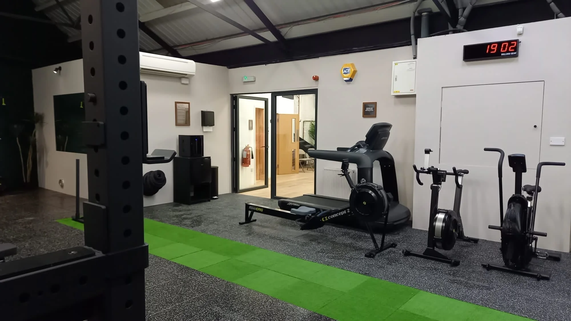 Gym interior with exercise machines, green floor strip, and digital clock. TRIBES St John's Wood, London NW8 0JT..