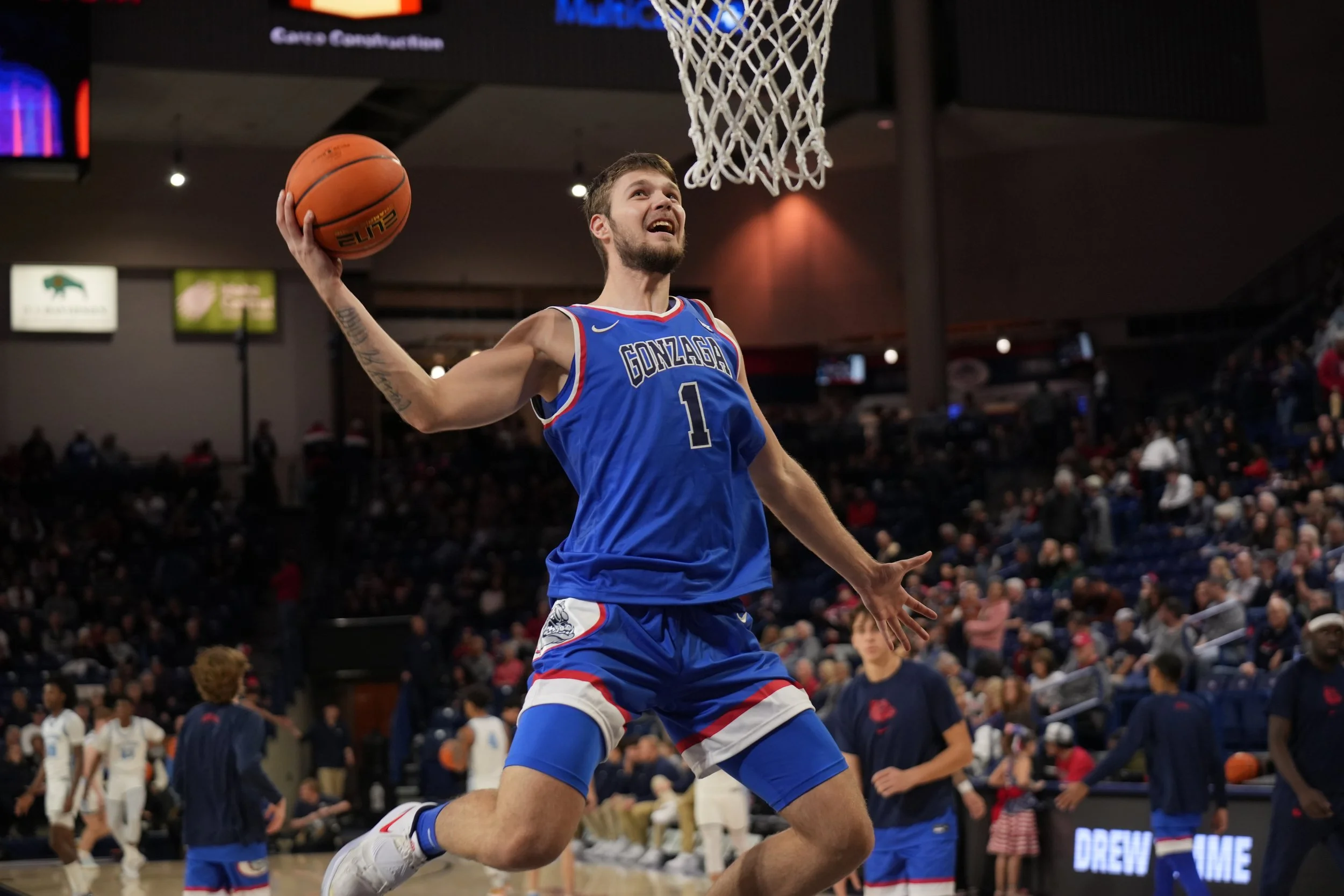Basketball player in blue Gonzaga jersey going for a slam dunk on a court during a game, with an audience in the background.