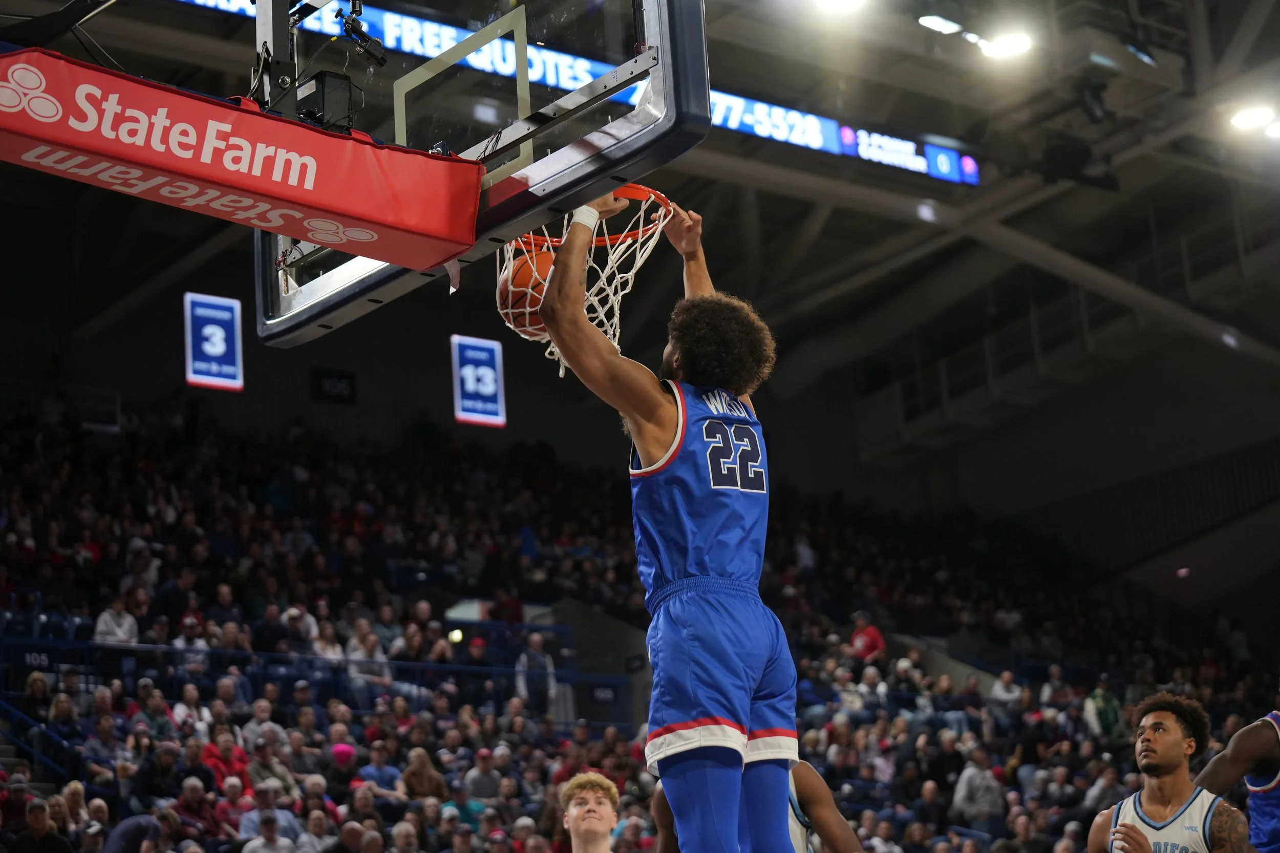 Basketball player in blue jersey dunking a basketball during a game, with spectators in the background.