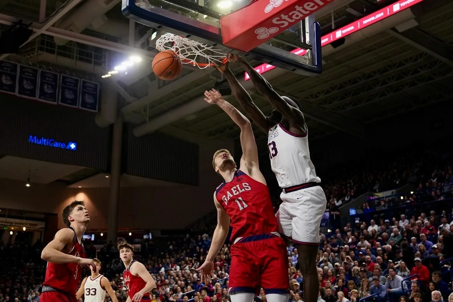 Basketball game action shot with a player in white jersey blocking a shot by a player in red jersey under the hoop, with spectators in the background.