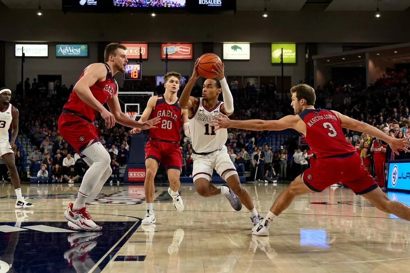 Basketball game action featuring players in red and white uniforms, with one player holding the ball while surrounded by defenders on a court.