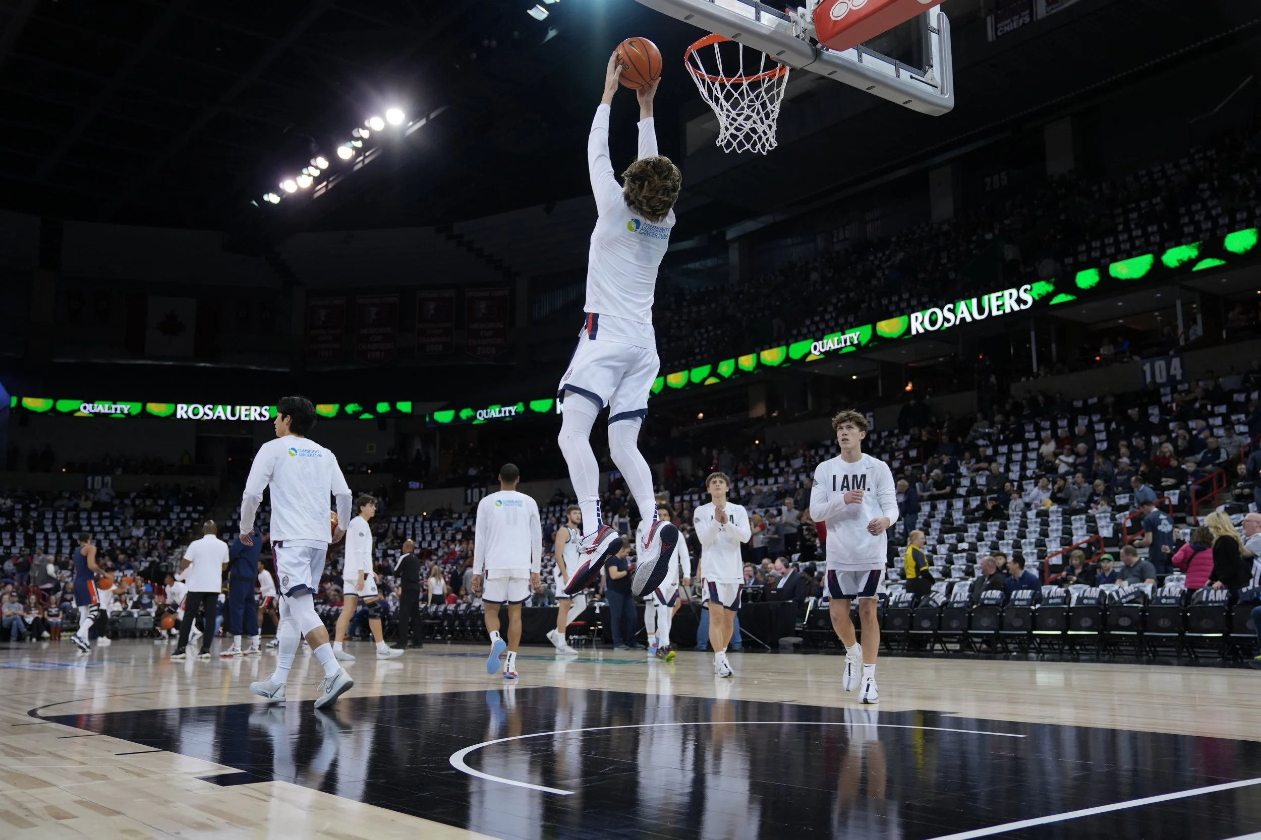 Basketball players warming up on a court, one player attempting a dunk. The stands are partially filled with spectators. The arena has advertisements visible.