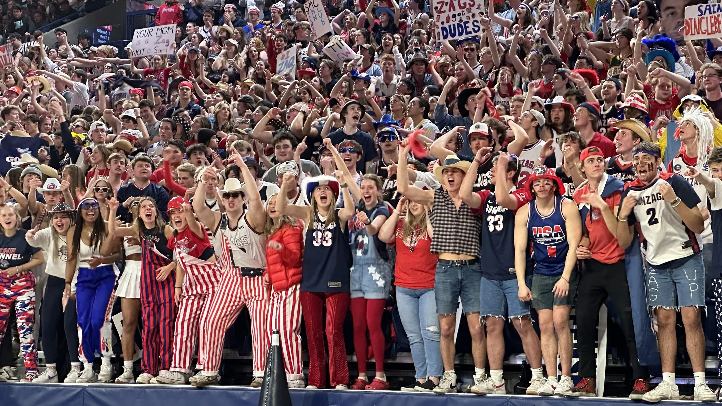 A large, enthusiastic group of sports fans cheering in a stadium, many wearing red, white, and blue attire and holding signs supporting Gonzaga University.
