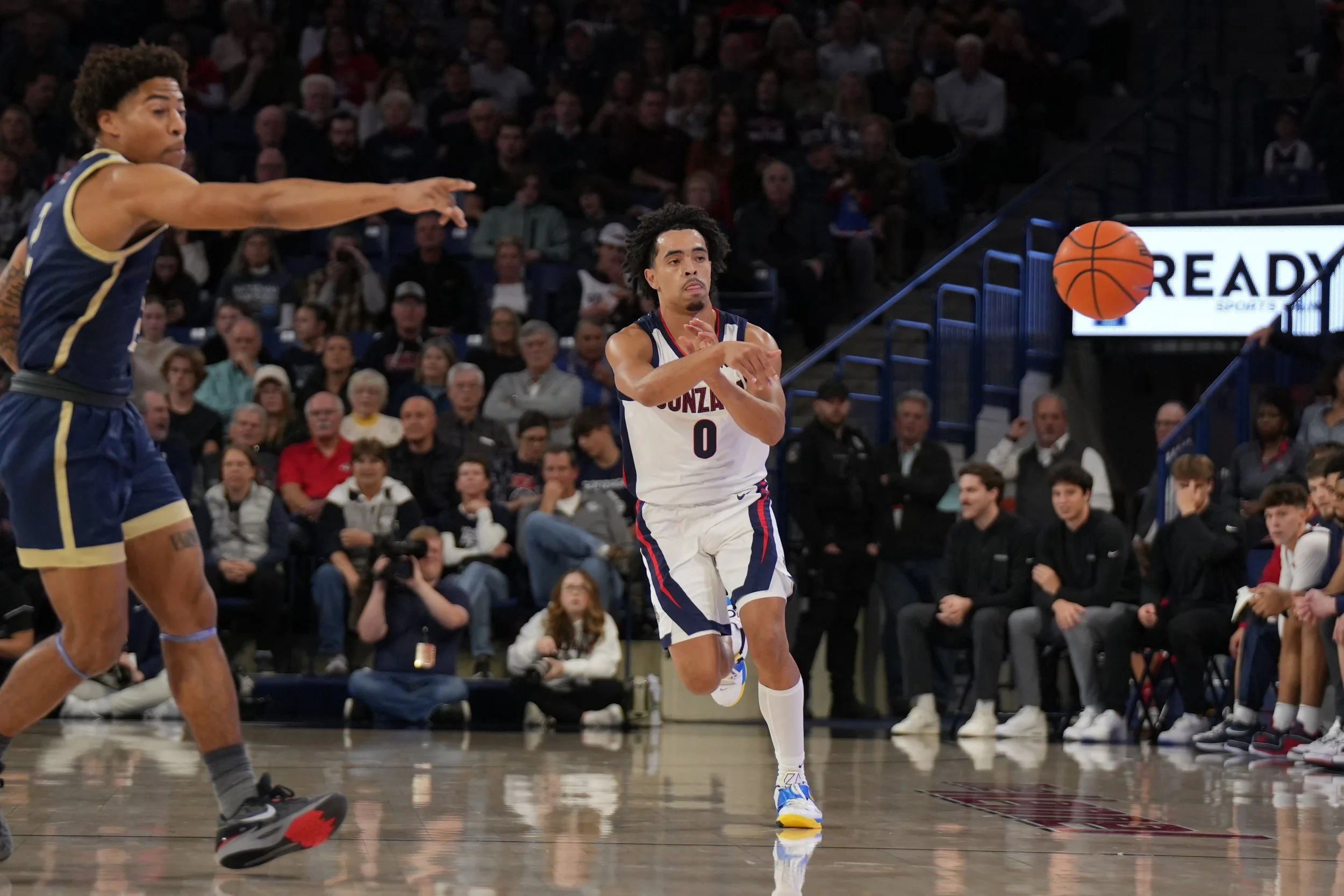 Basketball player in white uniform passing the ball during a game, with a defender in blue uniform attempting to block. Crowd in the background.