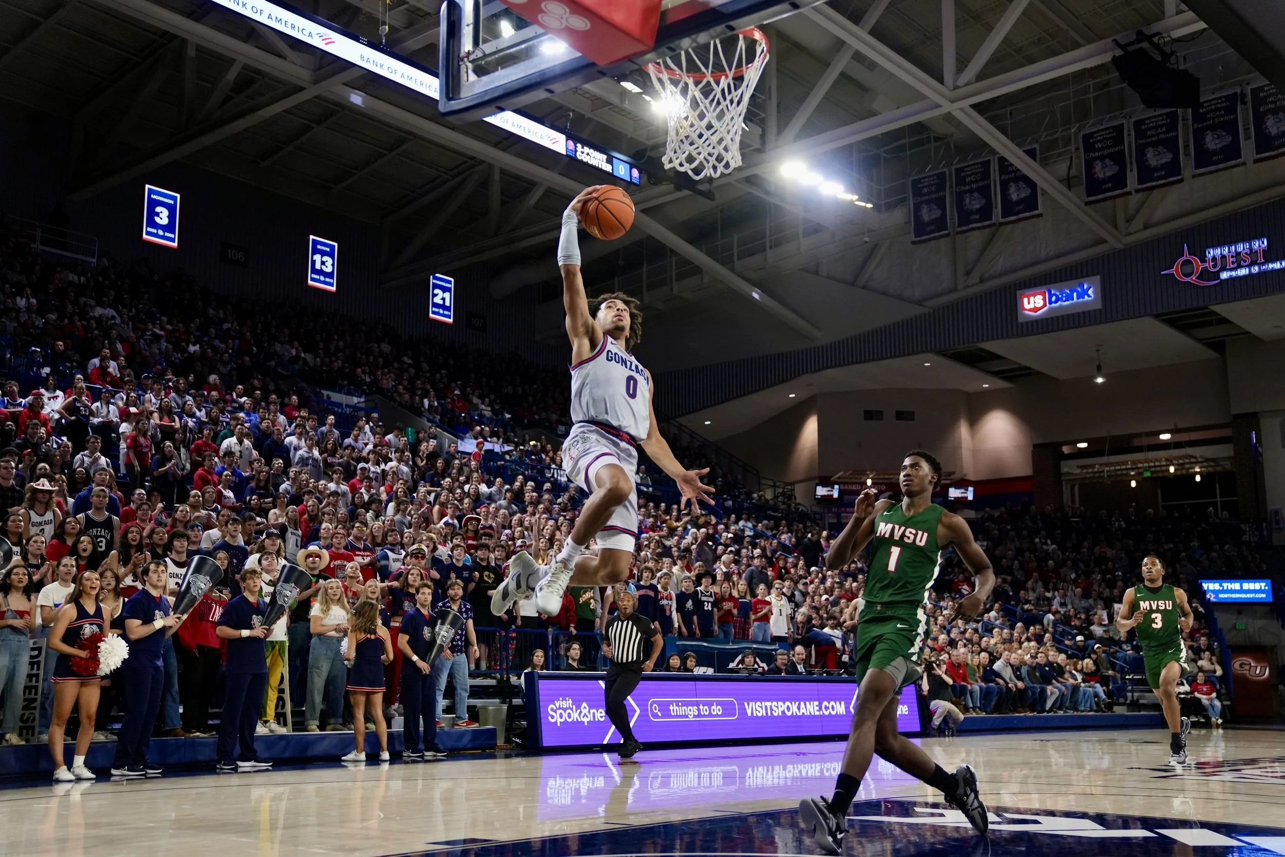 Basketball player in white jersey dunking during a game with a cheering crowd in the background. Opposing players in green jerseys. Indoor court setting.