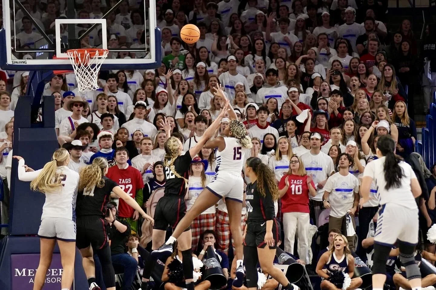 Women's basketball game with players from opposing teams jumping near the hoop. Crowd of fans in background wearing white shirts, some holding banners. Cheering cheerleaders visible.