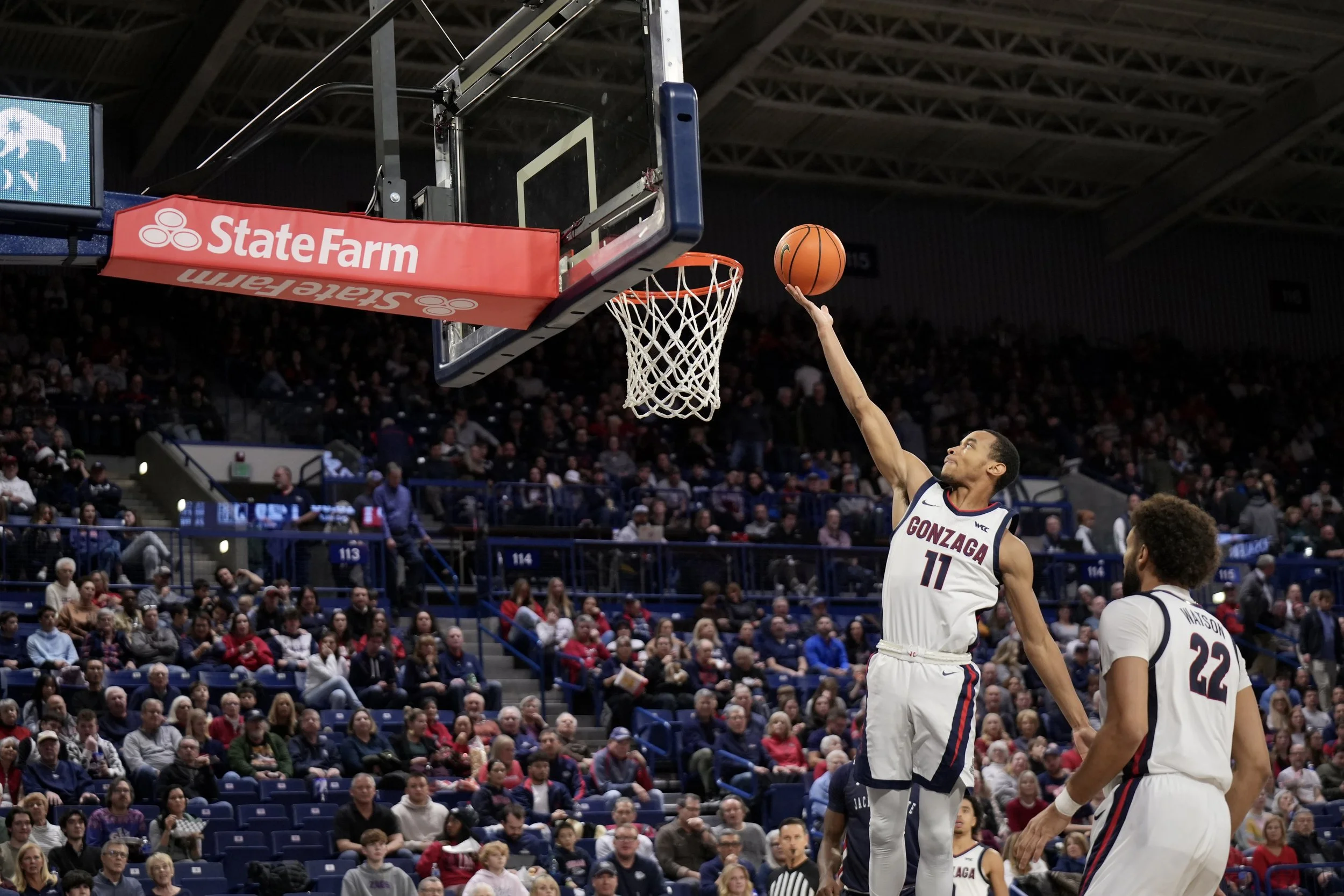 Basketball player in Gonzaga jersey making a layup during a game, with a crowd watching in the background.