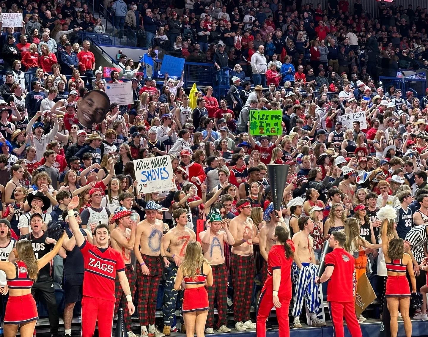 Crowd of enthusiastic fans at a sports event, many wearing red and blue. Some hold signs and others have body paint, with one large face cutout visible. Cheerleaders in red uniforms and people in costumes are interacting with the crowd, creating a li