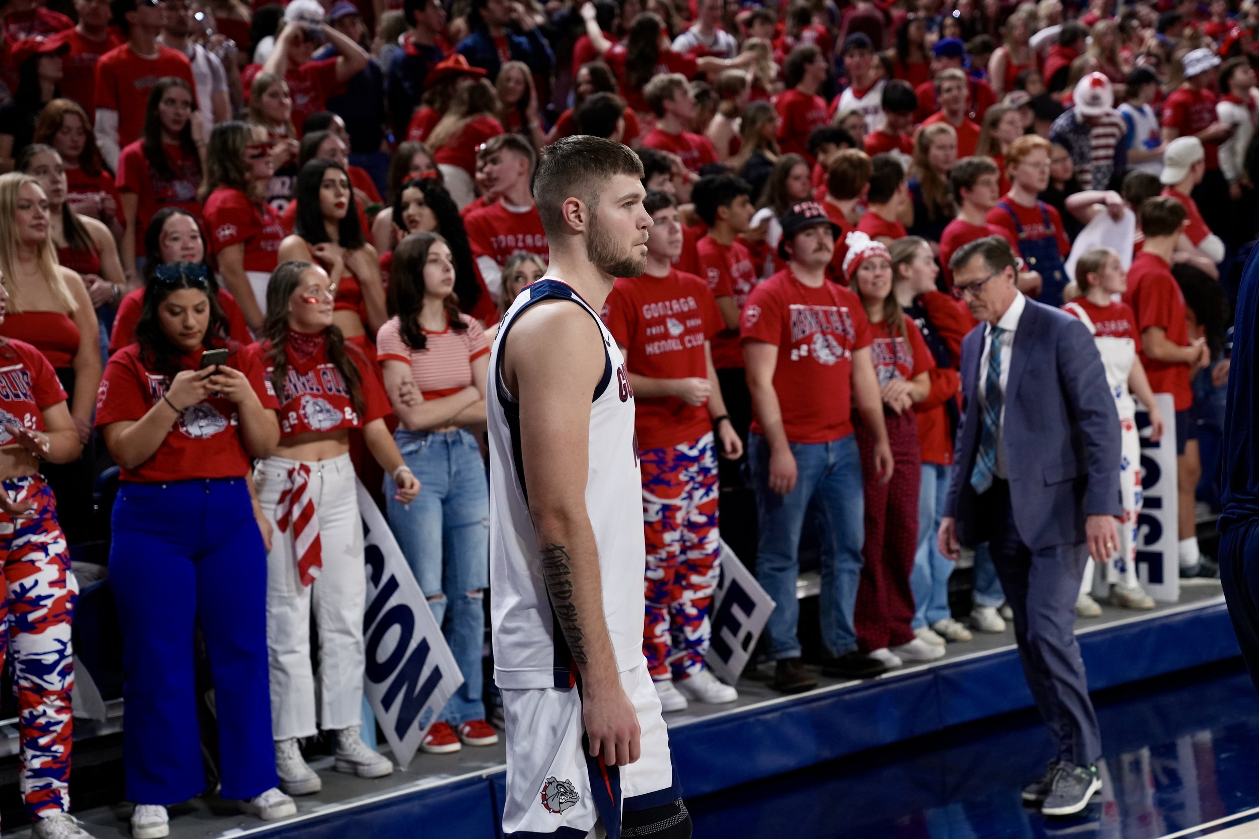 College basketball player on court surrounded by a cheering crowd in red attire, some holding banners.