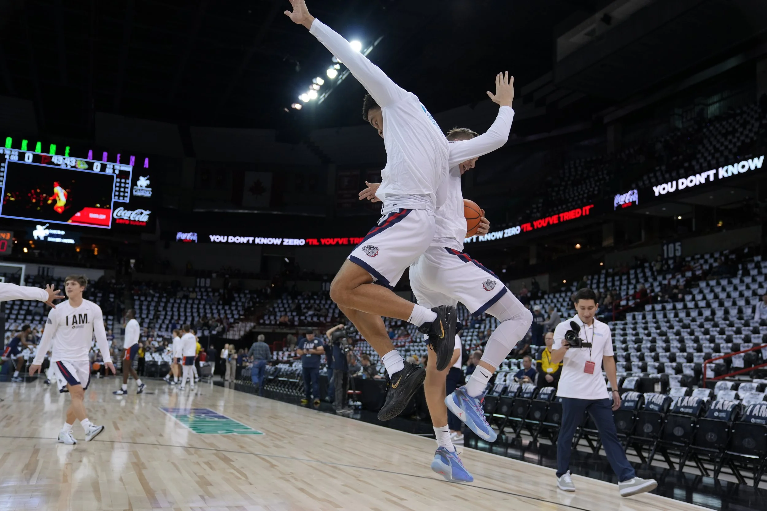 Basketball players on a court in a stadium, jumping in mid-air, surrounded by empty seats and a few people, including a photographer, in the background.