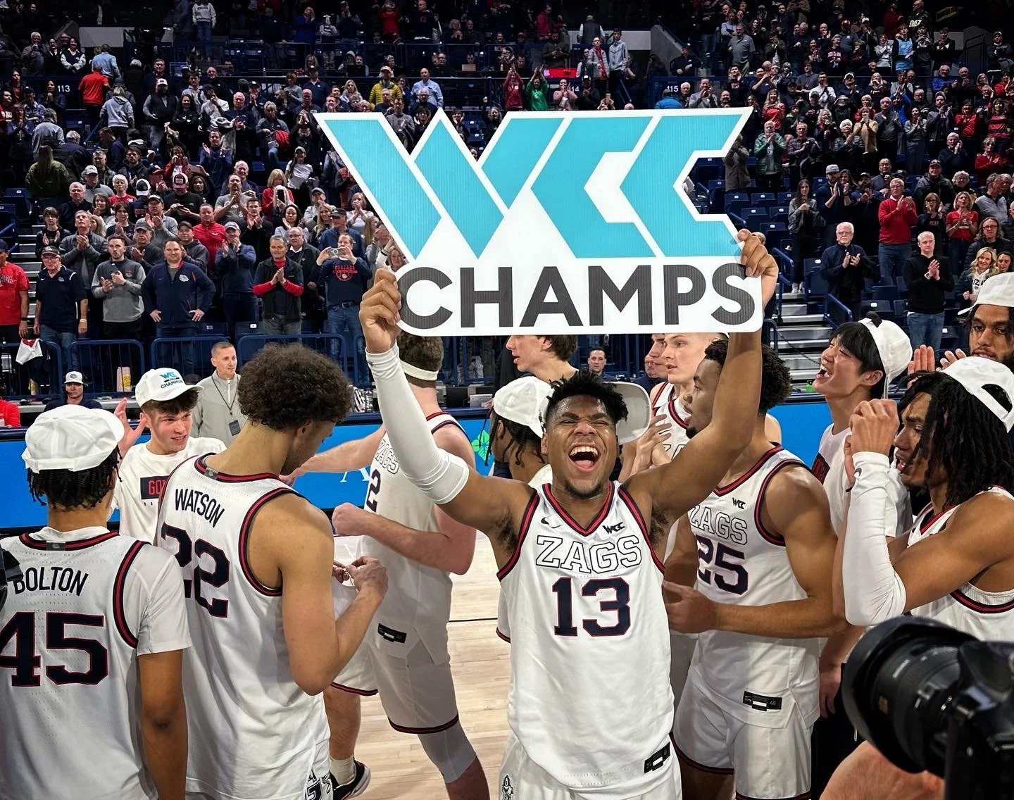 Basketball team celebrating WCC championship victory, holding "WCC Champs" sign on court, with cheering crowd in background.