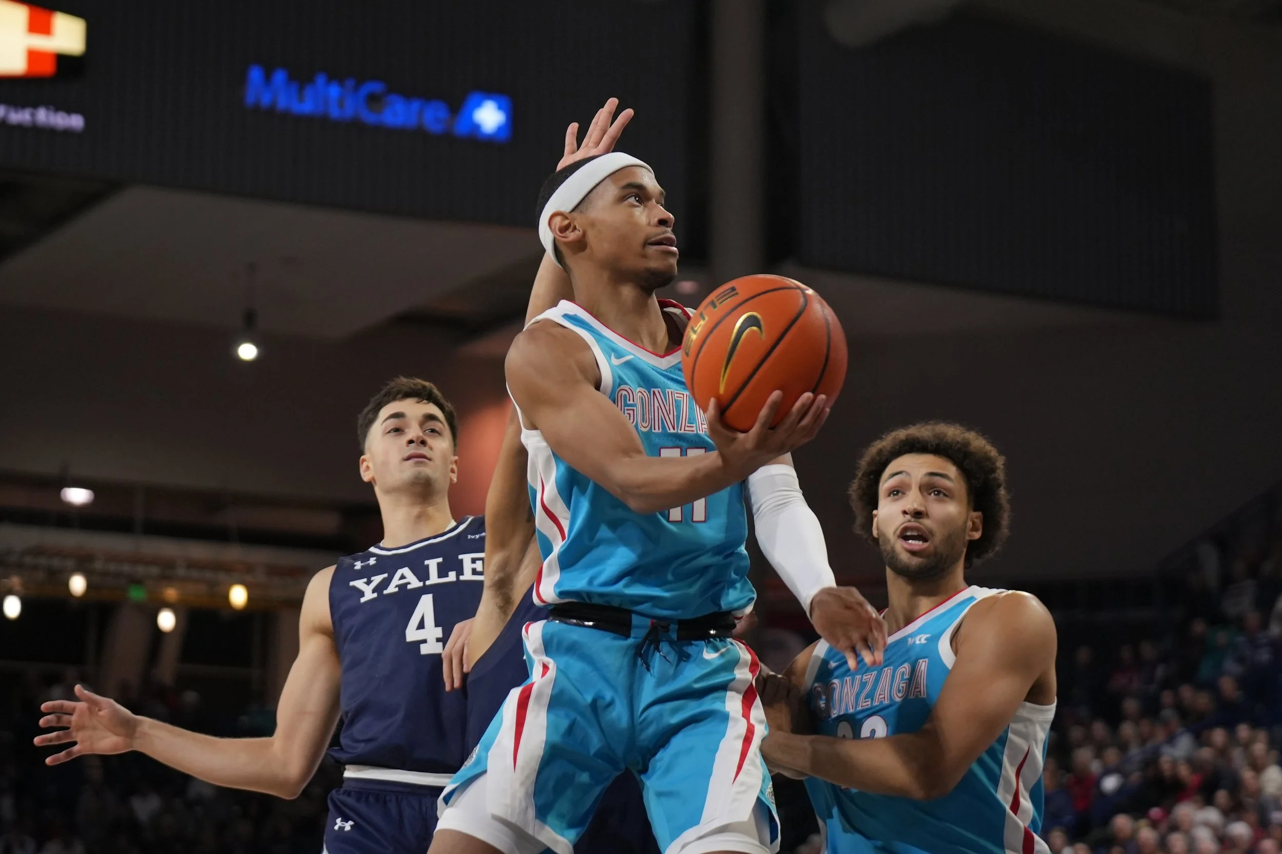 Basketball game with players from Gonzaga and Yale teams; Gonzaga player holding the ball, Yale player and another Gonzaga player nearby, court background.