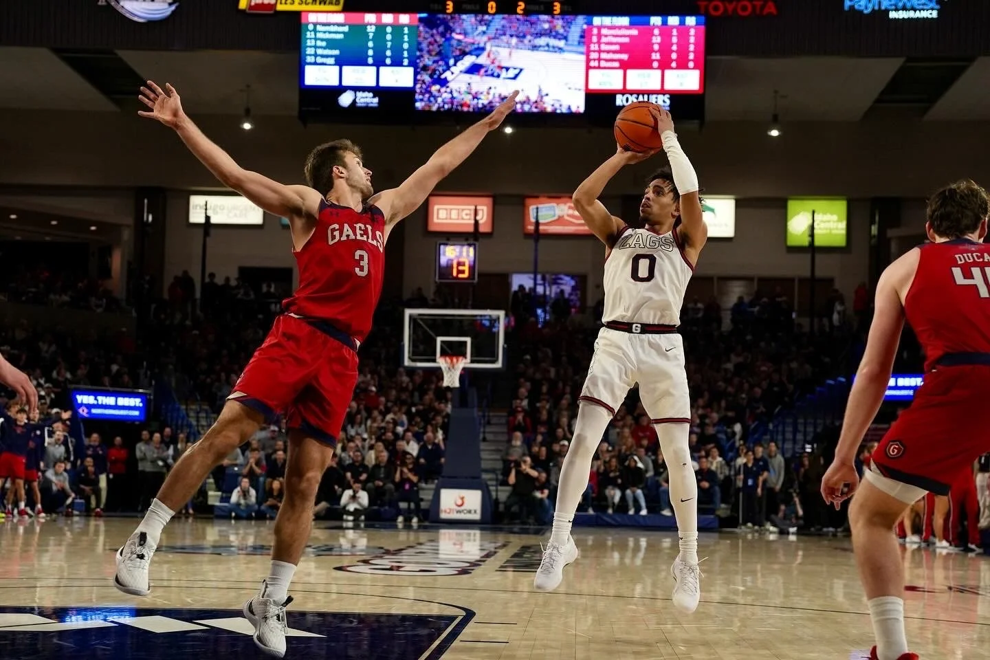 Basketball player in white jersey shooting over defender in red jersey during a game, with audience and scoreboard in background.